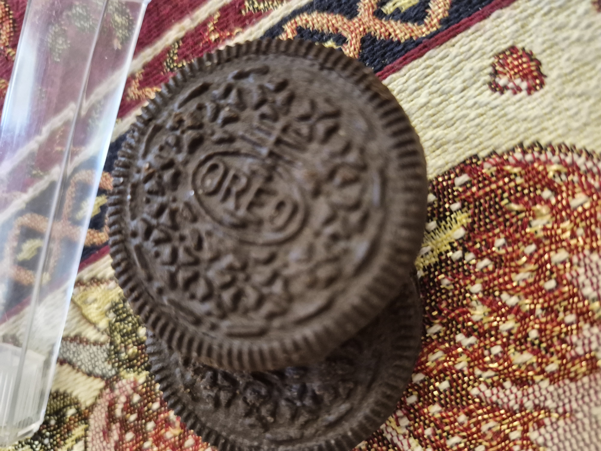 Two dark chocolate sandwich cookies, identifiable as Oreos by the embossed "OREO" text and distinctive pattern, are stacked on top of each other. The top cookie is in sharp focus, showcasing its intricate design and ridged edges. The cookies are positioned on a colorful, woven textile surface. This fabric features a patterned design with dominant colors of off-white/beige, red, dark blue/black, and yellow/gold, suggesting a traditional or ethnic style, potentially aligning with the cultural aesthetics found in Yerevan, Armenia. To the left of the cookies, a translucent, clear plastic object is partially visible, reflecting light and slightly obscuring a portion of the textile beneath it. The scene is a close-up, still-life arrangement, under what appears to be ambient indoor lighting. No specific activity or time of day is discernible, and no human subjects are present.