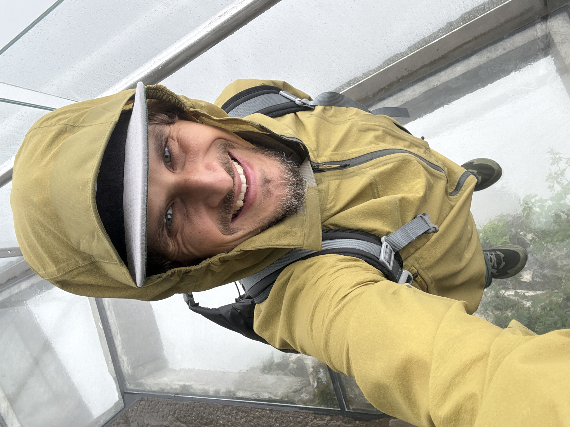 A man in a tan hooded jacket and baseball cap stands on a glass-bottomed walkway overlooking a mountainous landscape shrouded in fog. He is smiling and looking into the camera, which is held at an upward angle. The walkway appears to be wet and possibly outdoors. Based on the context, this location could be Tianmen Mountain in Yongding District, Zhangjiajie, China.