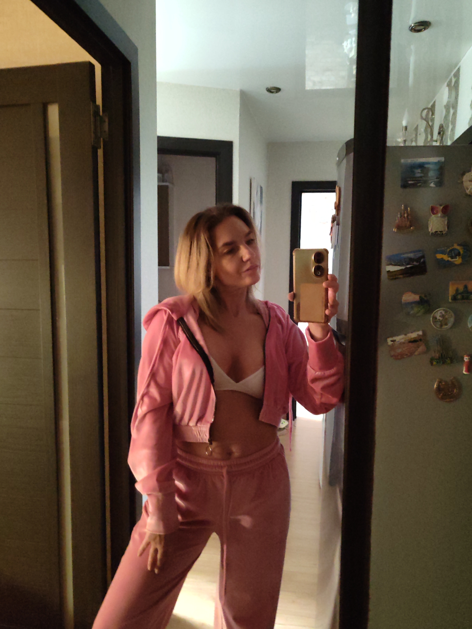 A woman with shoulder-length blonde hair is standing in what appears to be an apartment hallway. She is wearing a light pink velour tracksuit jacket and pants, with a white bra visible under the open jacket. She is holding a gold-colored smartphone, presumably taking a selfie in front of a mirror.
The hallway extends into the apartment, with doorways visible in the background. The mirror is attached to what seems to be a refrigerator covered in various magnets. The scene is indoors and well-lit. There is no evidence to suggest this photograph was taken in Petropavlovsk-Kamchatsky, Russia.