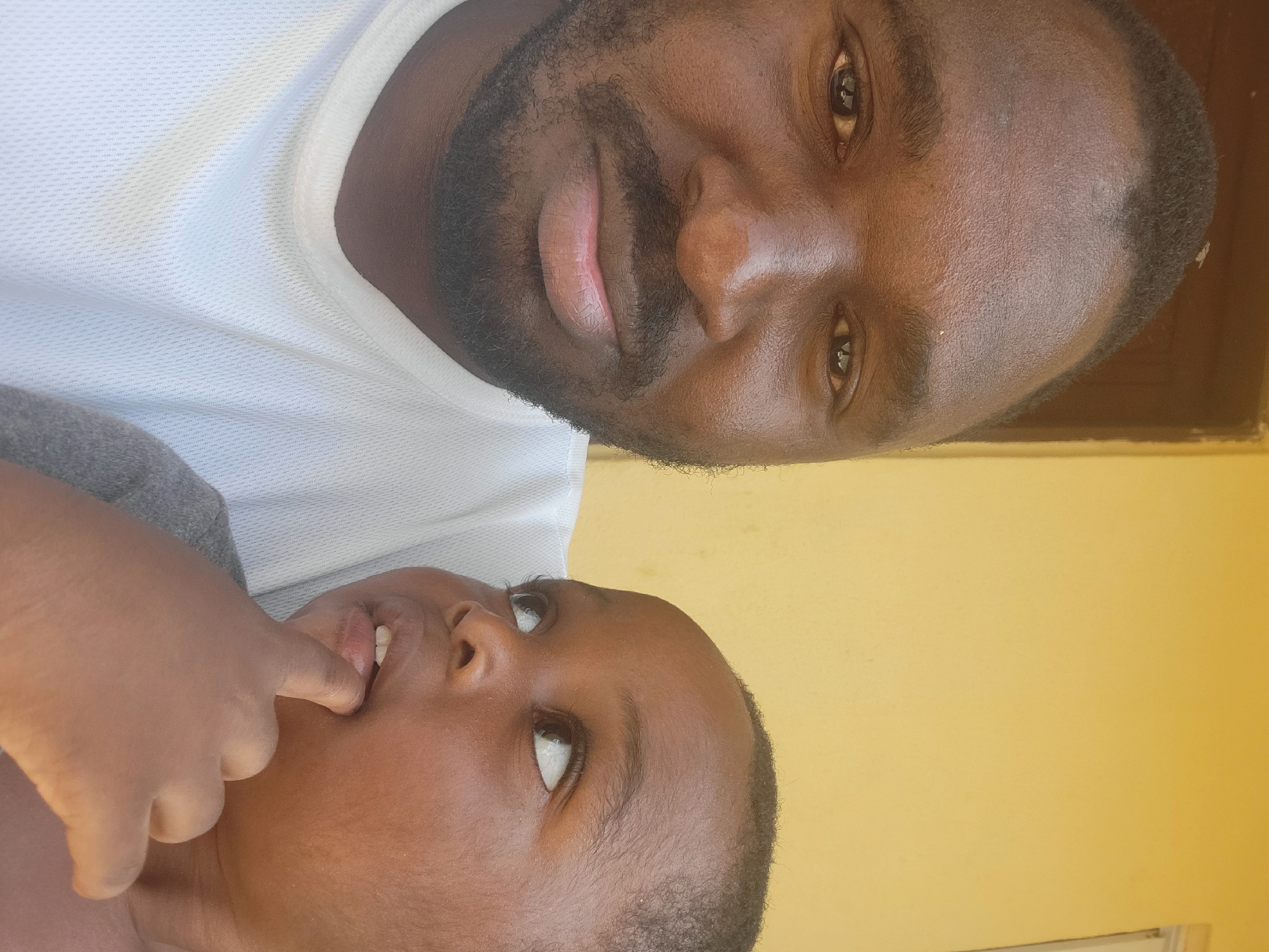 A close-up selfie shows a Black man in the foreground looking at the camera with a slight smile. He has dark skin, short black hair, a beard, and is wearing a white mesh t-shirt. To his right and slightly behind him, a young Black child is looking up and to the side, with their finger in their mouth. The child has dark skin and short black hair, and is wearing a gray shirt. The background is a plain yellow wall, with a sliver of a brown doorframe visible on the left. The lighting suggests it is daytime. The image appears to have been taken outdoors in Suleja, Nigeria.