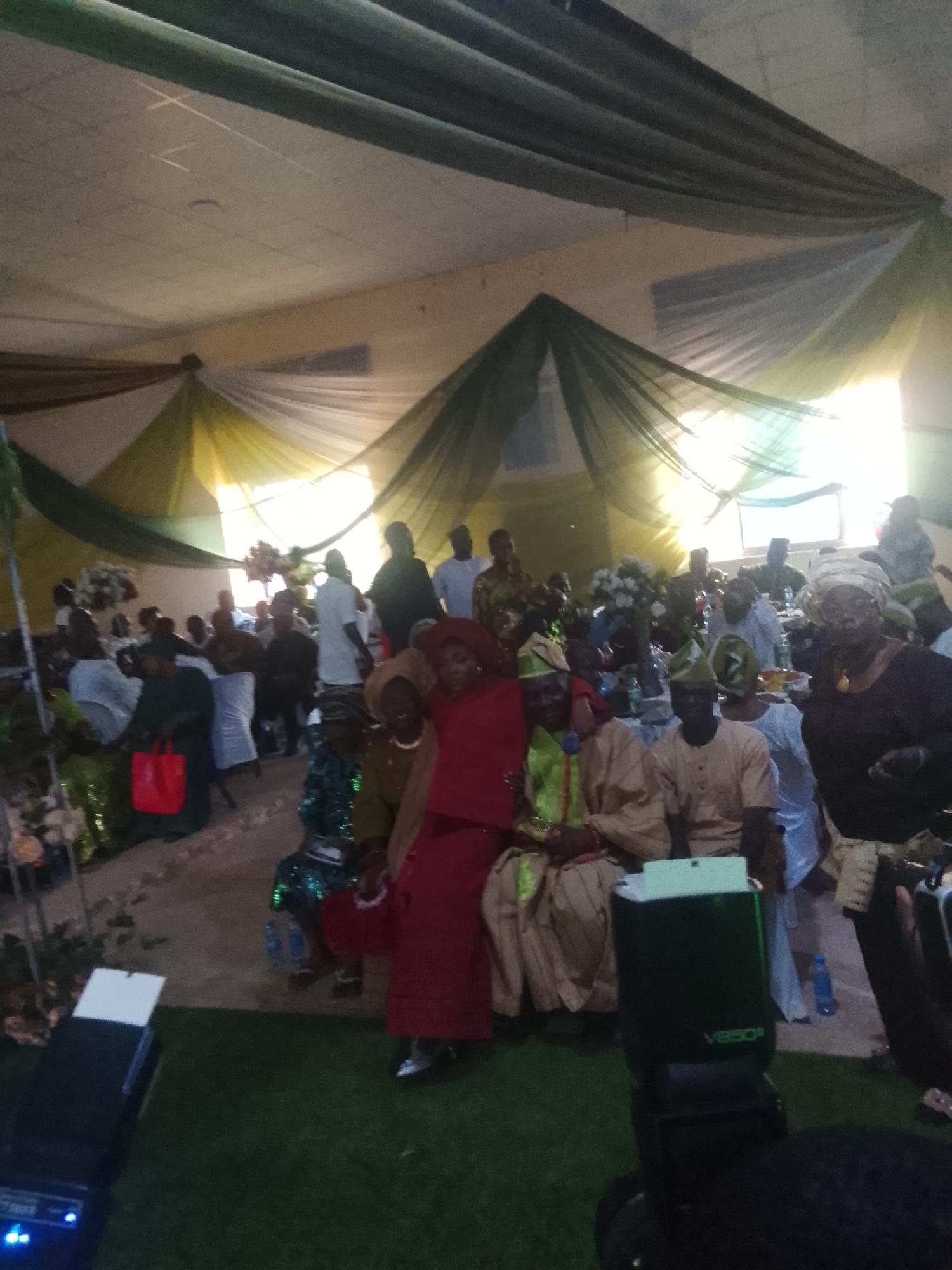 The image captures an indoor event with numerous people, likely a wedding or celebration, in a city of Unknown, Unknown. Green and white drapes adorn the ceiling, suggesting a decorated venue. Attendees are seated at tables, some eating. Many individuals wear traditional clothing. In the foreground, a camera with a "V850" flash is present.