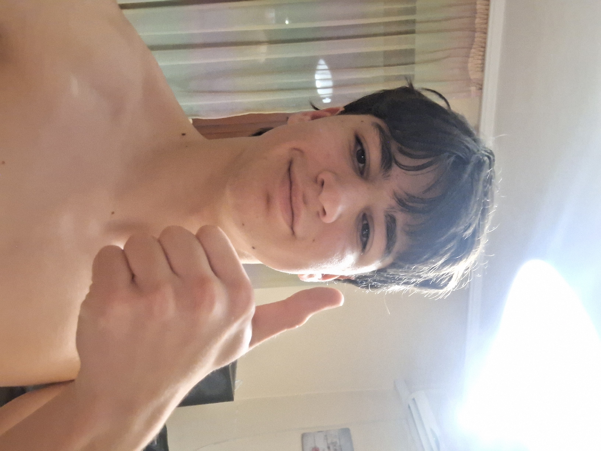 A bare-chested individual with dark hair is posing indoors, giving a thumbs-up. The location cannot be determined to be Burgas, Bulgaria from the image. There are blinds and a partial view of a wall.