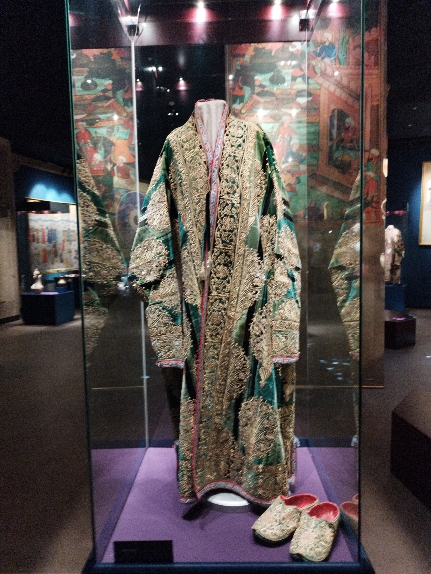 An ornate, emerald green and gold embroidered robe is displayed in a glass case. The robe has intricate gold detailing along its edges and appears to be made of a heavy fabric. Beneath the robe, on a purple platform, are two matching embroidered slippers. The background within the case features a colorful mural depicting figures in historical attire. The display case is situated in a museum setting, with other exhibits visible in the blurred background.