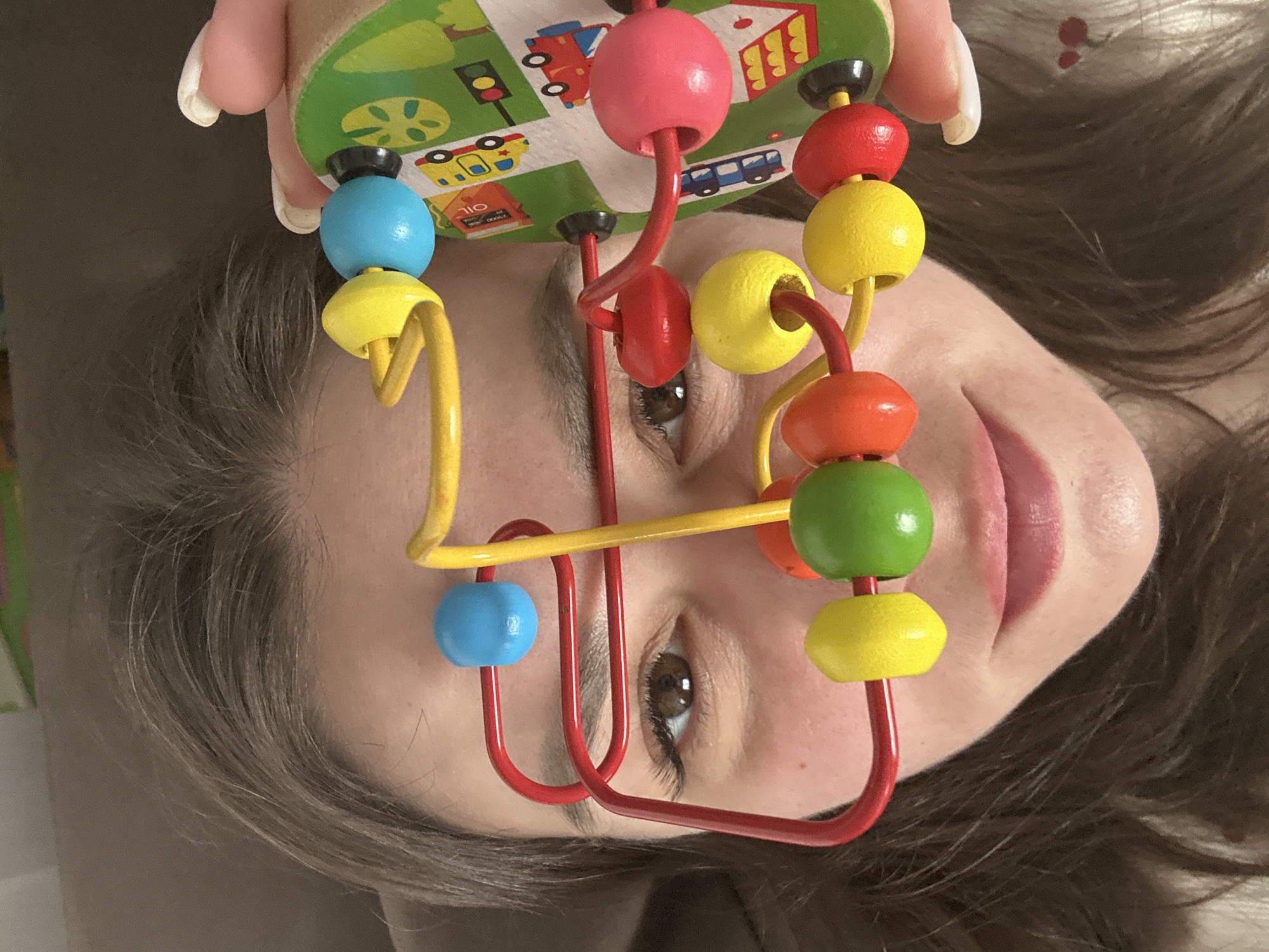 The image depicts a person's face, likely a woman, partially obscured by a colorful toy bead maze. The toy consists of wooden beads in shades of blue, red, yellow, green, and orange threaded on red and yellow metal wires, which are attached to a green and black base with vehicle graphics. The person is holding the toy, placing it in front of her face. The photograph appears to have been taken indoors with natural lighting. No information is available to confirm that the image was taken in Chaikovsky, Russia.