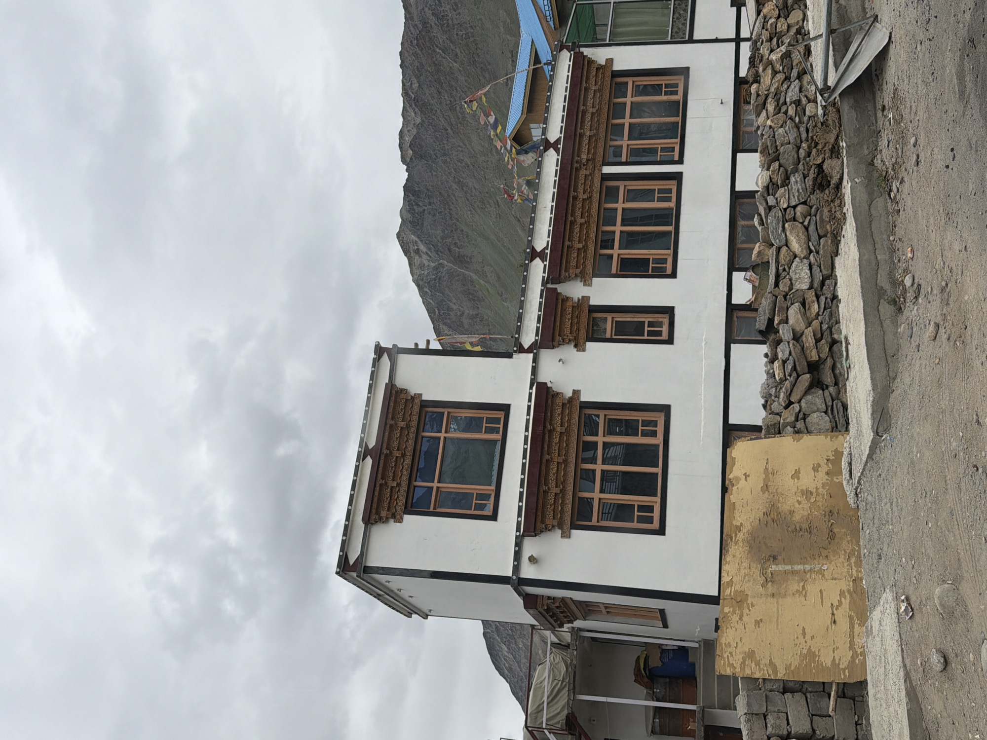 The image shows a two-story white building with dark trim and ornate wooden window frames, located in Padum, India. The building has several windows on both floors, some with multiple panes. Prayer flags are strung across the roofline, and behind the building, a rocky mountain looms under a cloudy sky. In front of the building, there is a low wall constructed of rough stones, and a large, weathered piece of plywood leans against it. The ground in front of the wall is a mix of dirt and gravel, with some concrete slabs. There are no people visible in the image. The scene appears to be a rural or mountainous area, consistent with the location of Padum.