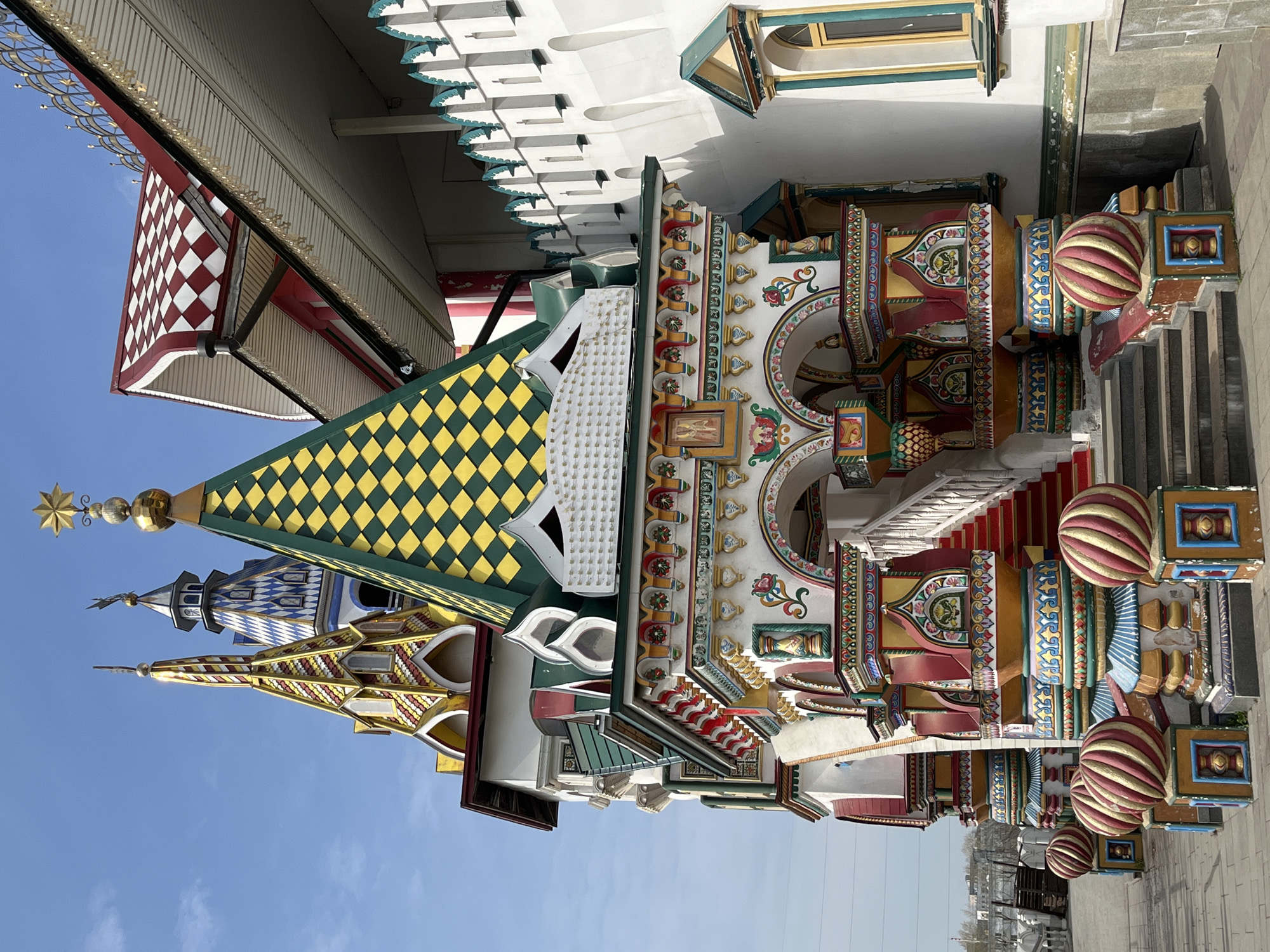 The image shows the colorful and ornate architecture of St. Basil's Cathedral in Moscow, Russia. The cathedral features multiple domes and towers with unique patterns and colors. The sky is visible in the background, indicating it is daytime. The building's details include geometric patterns, arches, and decorative elements. The foreground shows a pedestrian walkway, suggesting the building is accessible to the public.