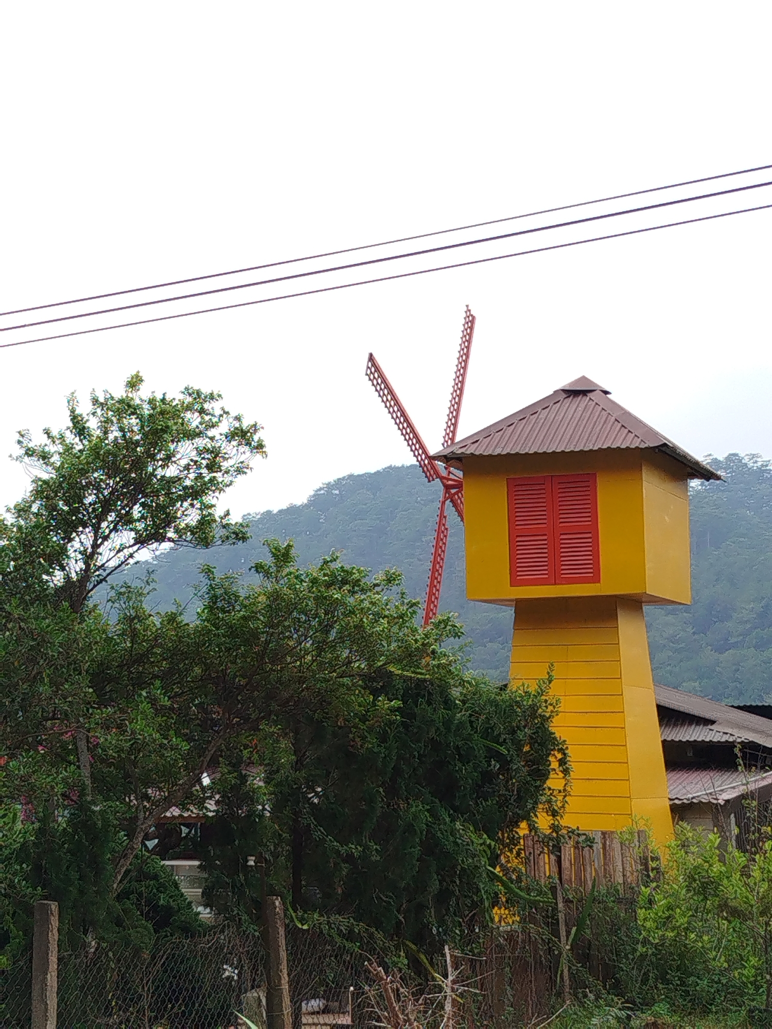 The image shows a brightly colored yellow windmill-like structure with red accents in Da Lat, Vietnam. The structure is partially obscured by lush green foliage. It has a brown, peaked roof and a square upper section with red shuttered windows. The base of the structure is a yellow tower with vertical lines, resembling a stylized windmill. In the background, a red windmill is visible against a backdrop of a misty, green, tree-covered hill or mountain. Utility wires stretch across the upper portion of the frame. The foreground features a fence and more greenery, suggesting a rural or semi-rural setting within the city.