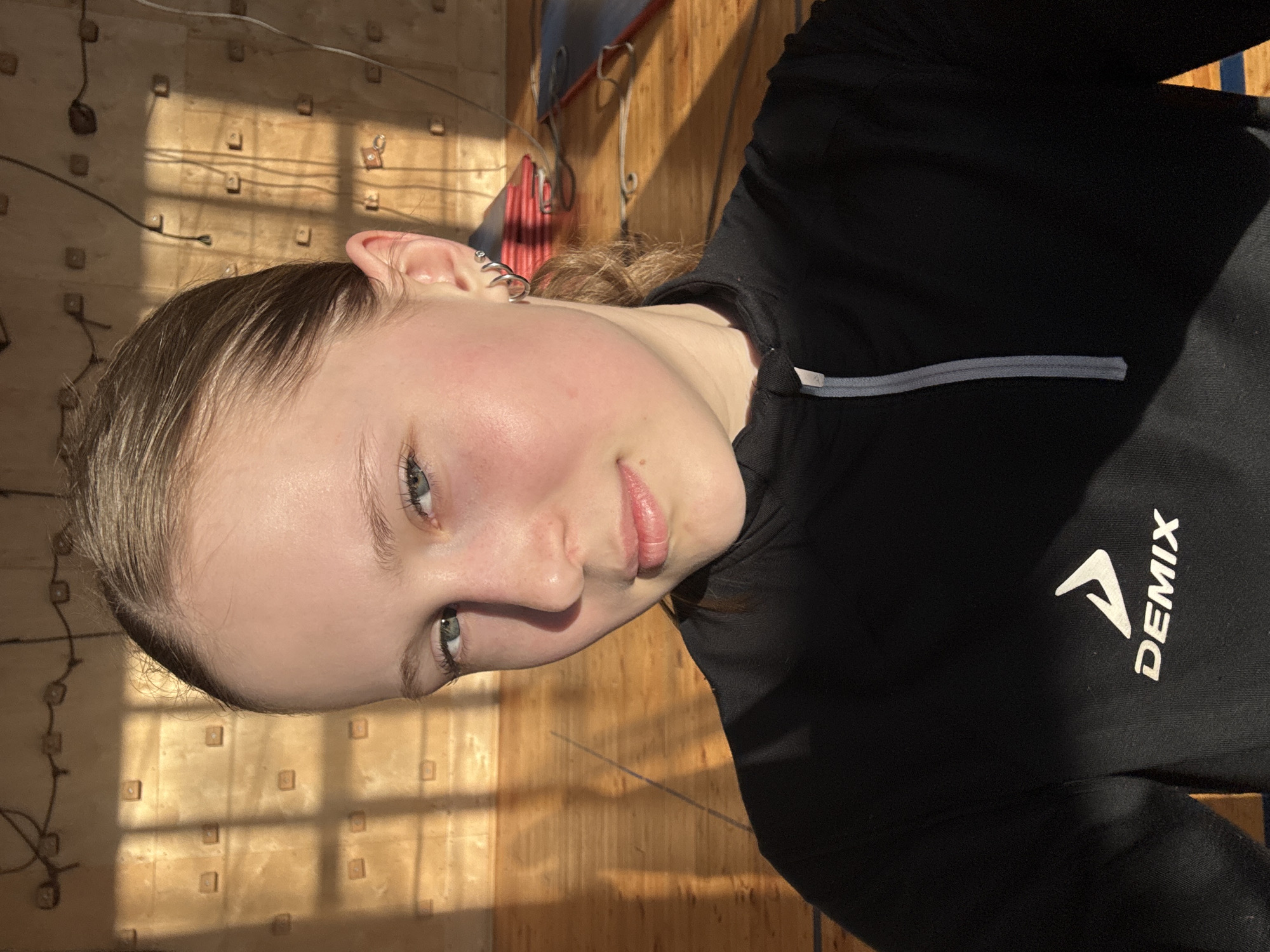 A close-up indoor shot features a young, fair-skinned person with light brown hair pulled back. The person faces the camera with a slight tilt, revealing light blue eyes. The person is wearing a black zippered athletic jacket with a white "DEMIX" logo visible on the right chest area. The background consists of a wooden wall, partially illuminated by sunlight creating a pattern of light and shadow resembling a grid. A red and grey object is partially visible in the background near the upper center. Given the request for specificity and the presence of the Demix logo, a clothing brand with a store in Chaikovsky, Russia, it is plausible that the location is in or near the city of Chaikovsky, Russia.