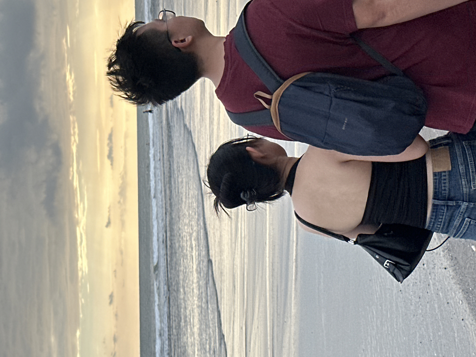 The image depicts two individuals, a woman and a man, from the back as they stand on a beach. The woman, in the foreground on the left, wears a black tube top and blue jeans, carrying a black handbag over her left shoulder. Her hair is tied up in a bun. The man, to her right and slightly further back, wears a burgundy t-shirt and glasses, with a dark blue backpack on his back. They are both facing the ocean, which is calm with gentle waves. The sky above is overcast with patches of yellow and orange light, suggesting either sunrise or sunset. The beach is wet and sandy, with the tide lapping at the shore. In the distance, a few figures can be seen in the water. The location is identified as the beach in North Kuta, Indonesia, a popular coastal area known for its beaches and sunsets.
