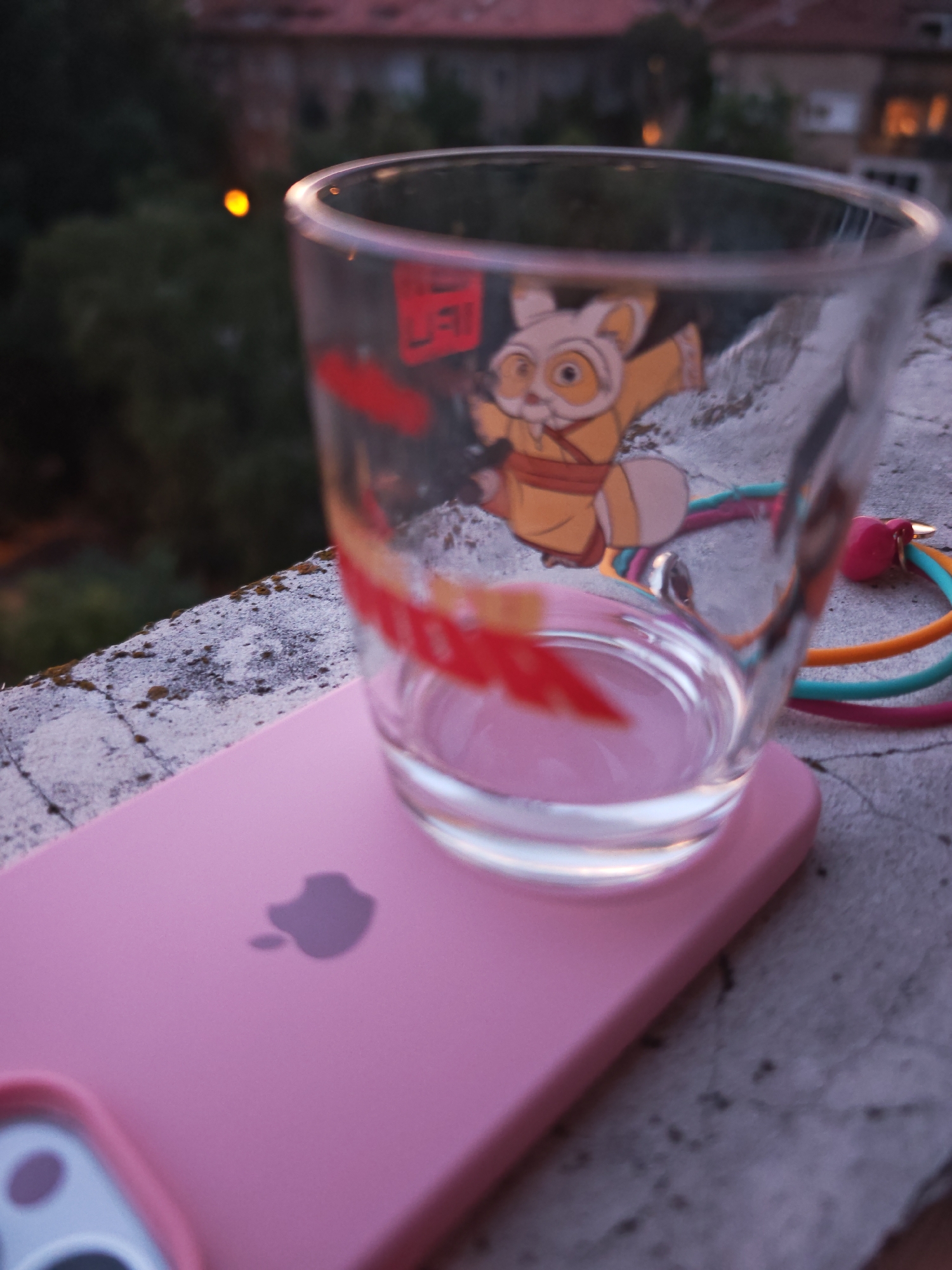 A close-up, eye-level shot captures a pink iPhone case lying flat on a textured, weathered concrete surface. Resting on the iPhone case is a clear glass with a character from Kung Fu Panda printed on it. The glass is transparent and partially filled with liquid. Several colorful, thin silicone cords, possibly charging cables or a lanyard, are visible extending from the side of the glass. The background is softly blurred, showing dark green foliage and hints of distant buildings with illuminated windows, suggesting an urban setting at dusk or dawn. The scene appears to be outdoors, possibly on a balcony or windowsill, in Sofia, Bulgaria, given the architectural style and vegetation visible in the background.