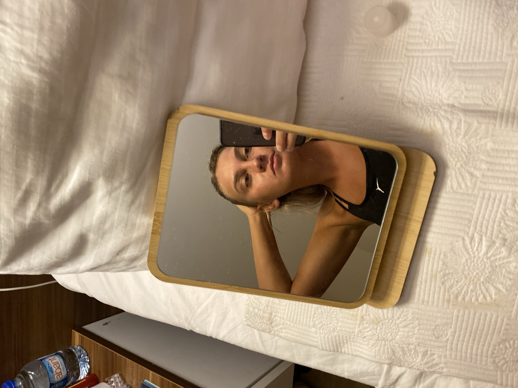In a bedroom, likely in Aksu, Turkey, a young woman is taking a selfie in a handheld mirror. She is wearing a black sports bra and has her hair pulled back. The mirror is propped up on a wooden stand on a white bedspread. In the background, a water bottle is visible on a nightstand, and a white pillow is propped up on the bed. The overall lighting is dim, suggesting it may be evening or nighttime.