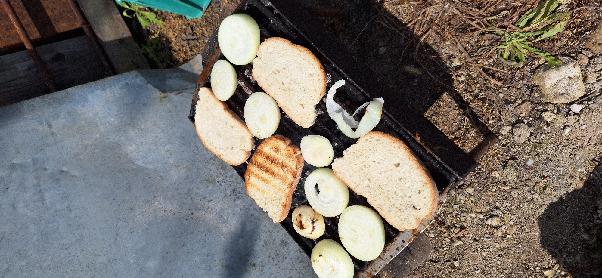 A slightly high-angle, outdoor shot shows a grill grate with various slices of bread and rings of onions. The grill sits on a textured metal surface to the left, and a dirt ground with small stones and plants to the right. Some bread slices are grilled, while others are light in color. The onion slices range from white to translucent. The location is Katuntsi, Bulgaria.