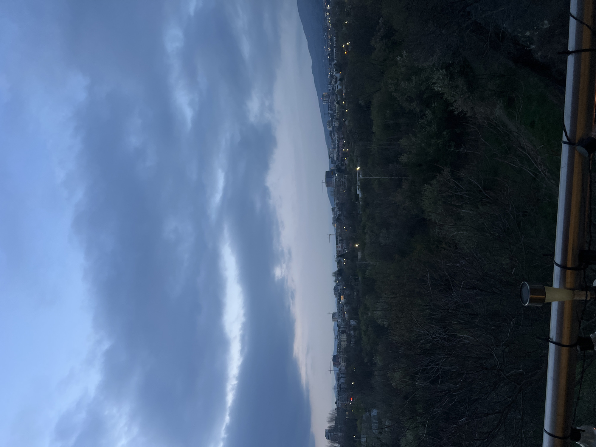 The image presents an evening scene likely from a balcony or elevated position overlooking part of Sofia, Bulgaria. The sky is predominantly cloudy, with varying shades of blue and grey. A mountain range is visible in the background. A line of buildings with streetlights is discernible along the base of the mountains, suggesting urban or suburban development. The foreground includes trees and vegetation, some appearing dark due to the low light conditions. Parts of a railing are present in the bottom right corner of the image.