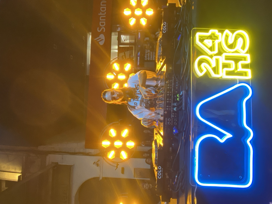 An outdoor DJ event is depicted at night in Buenos Aires, Argentina. A person with long, dark hair, wearing a white t-shirt with a graphic text including "NO TEARS," stands centrally, operating a professional DJ console. Headphones rest around their neck, and both hands are engaged with the mixer and media players. The DJ equipment bears the branding "COLO EQUIPOS." Behind the DJ, several hexagonal LED stage lights emit intense yellow-orange illumination. In the immediate foreground, a blue neon sign spells "BA" to the left, and a yellow neon sign spells "24 HS" to the right, mounted on a dark surface. The background reveals an urban environment: to the left, an older building with arched architectural elements is visible; to the right, a Santander bank branch is identifiable by its logo and a banner partially visible with Spanish text "Cobra y paga desde la Santand... Empresas." Several individuals are visible in the dimly lit background, observing the performance.