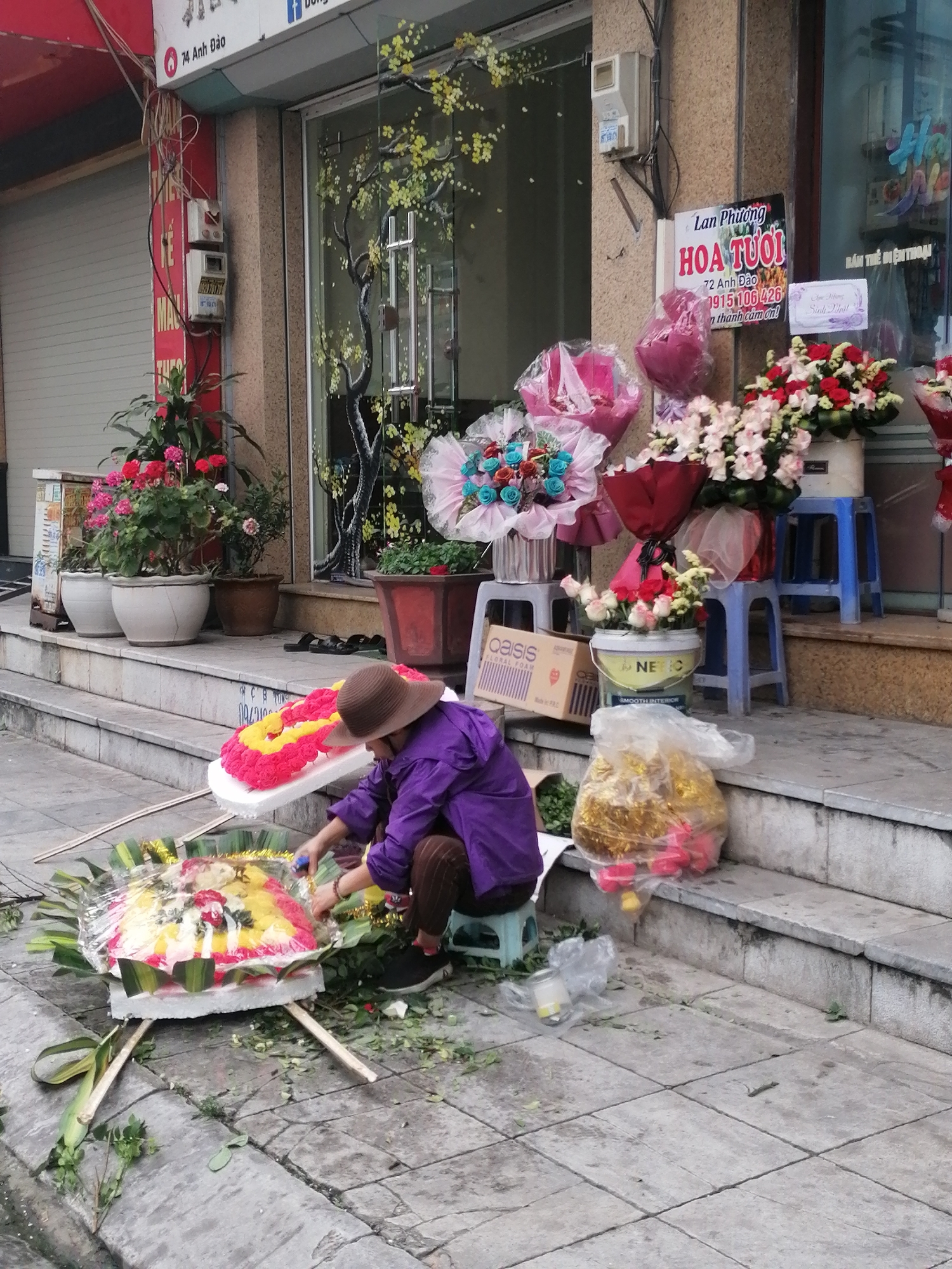 A street scene in Bai Chay Ward, Vietnam shows a woman seated on a small plastic stool, working on a floral arrangement on the sidewalk in front of a flower shop. She wears a brown hat and a purple jacket. She is creating a circular wreath with red, yellow, and white flowers, surrounded by large green leaves.
The flower shop has potted plants on the steps and bouquets displayed on stools. A sign reads "Lan Phương HOA TUOI" (Lan Phuong Fresh Flowers). Other visible details include an electrical meter, a storefront with decorative window art, bags of flowers, and floral foam packaging. The sidewalk is made of gray concrete.