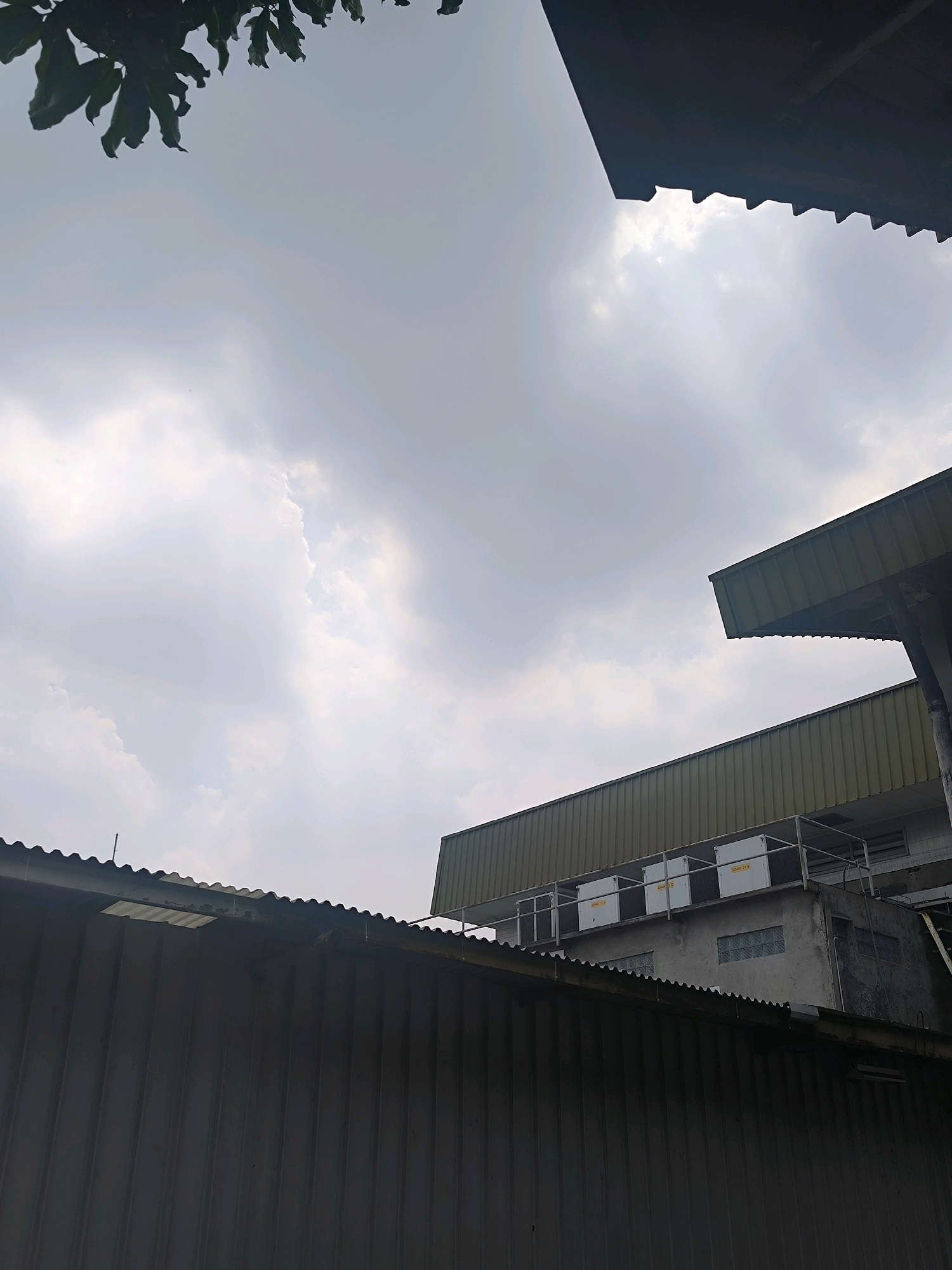 The image captures a cloudy sky over an industrial area in Bandung City, Indonesia. The foreground is dominated by the corrugated metal roofs of two adjacent industrial buildings, the one in the foreground being a dark gray, and the one behind it a muted olive green. The buildings appear to be multi-story structures with exposed ventilation units and pipes visible on the upper levels. The sky is overcast with diffused sunlight breaking through the clouds, creating a soft, diffused illumination. In the top left corner, a cluster of dark green leaves from a nearby tree hangs into the frame, providing a contrast to the man-made structures. The overall impression is one of an industrial landscape under a gray, atmospheric sky.