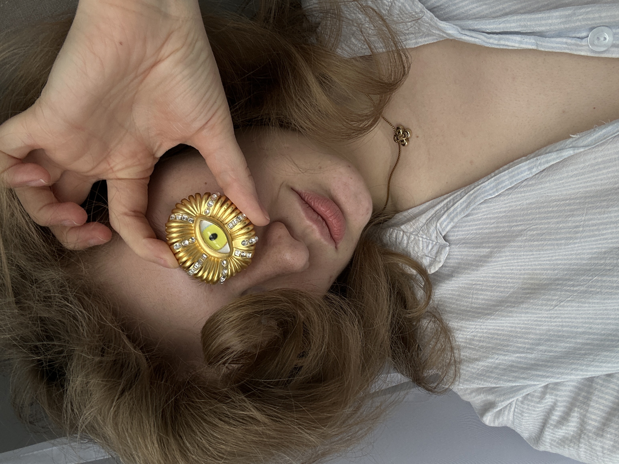 A person is reclining and facing upward. Their hand holds a golden brooch with an eye design near their face. They are wearing a light blue striped shirt. A gold necklace with a floral pendant is visible on their chest. The location cannot be determined.