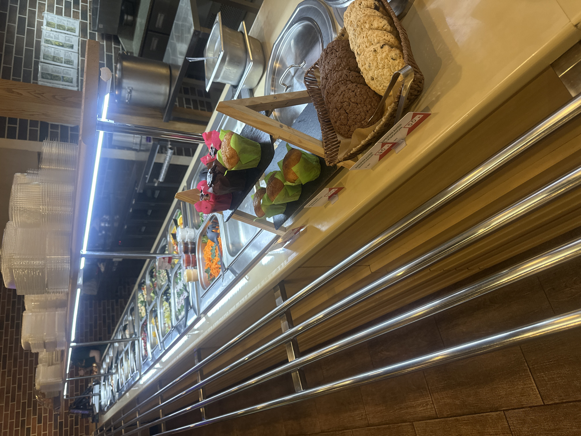 The image shows a food service area in Krasnaya Polyana, Russia, possibly a buffet or salad bar within a restaurant or cafeteria setting. On display are assorted salads and toppings, alongside wrapped muffins and a basket of cookies/biscuits. There is a price tag on the muffins, reading 50. The background reveals additional kitchen equipment such as a large cooking pot and stainless steel containers. Stacks of clear plastic cups are visible on the left. The scene is well-lit, with lighting fixtures visible above. The foreground features horizontal metal bars, possibly part of the serving station.