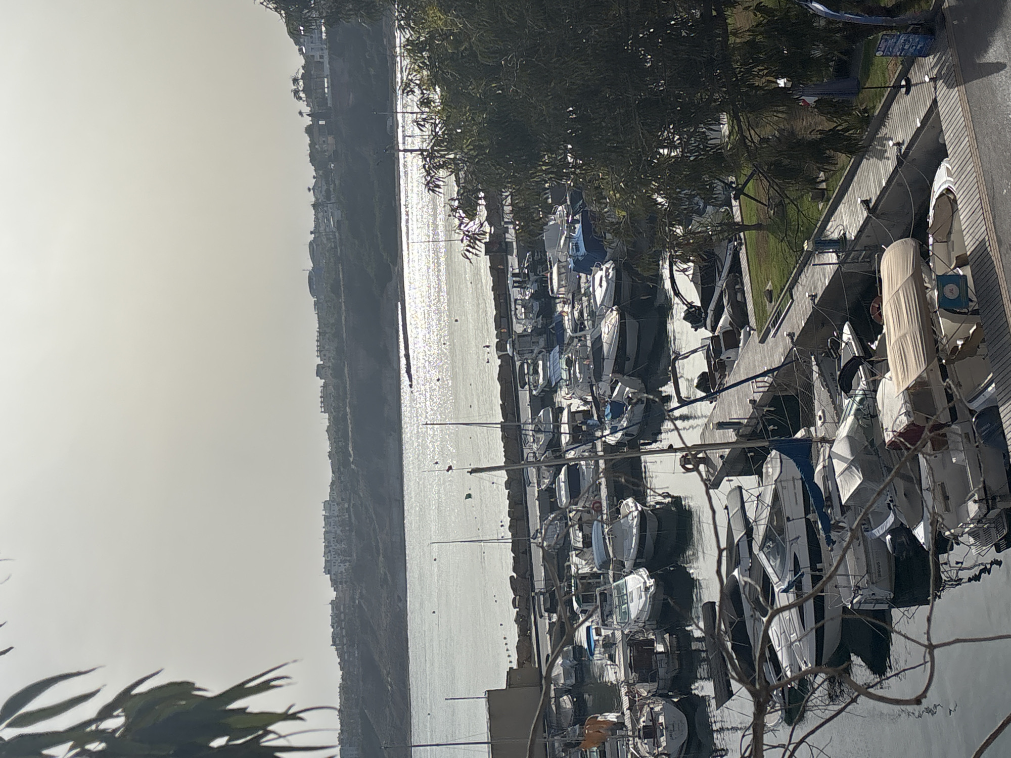 The image shows a marina with numerous boats docked. The boats vary in size and type, including sailboats and motorboats, many of which are covered with tarps. A wooden walkway runs alongside the docked boats. In the background, a body of water reflects the sunlight, and beyond that, a coastline with buildings and vegetation is visible. The sky is overcast with hazy sunlight. The scene is located in the city of Urbanización Cabo Roig, Spain.
