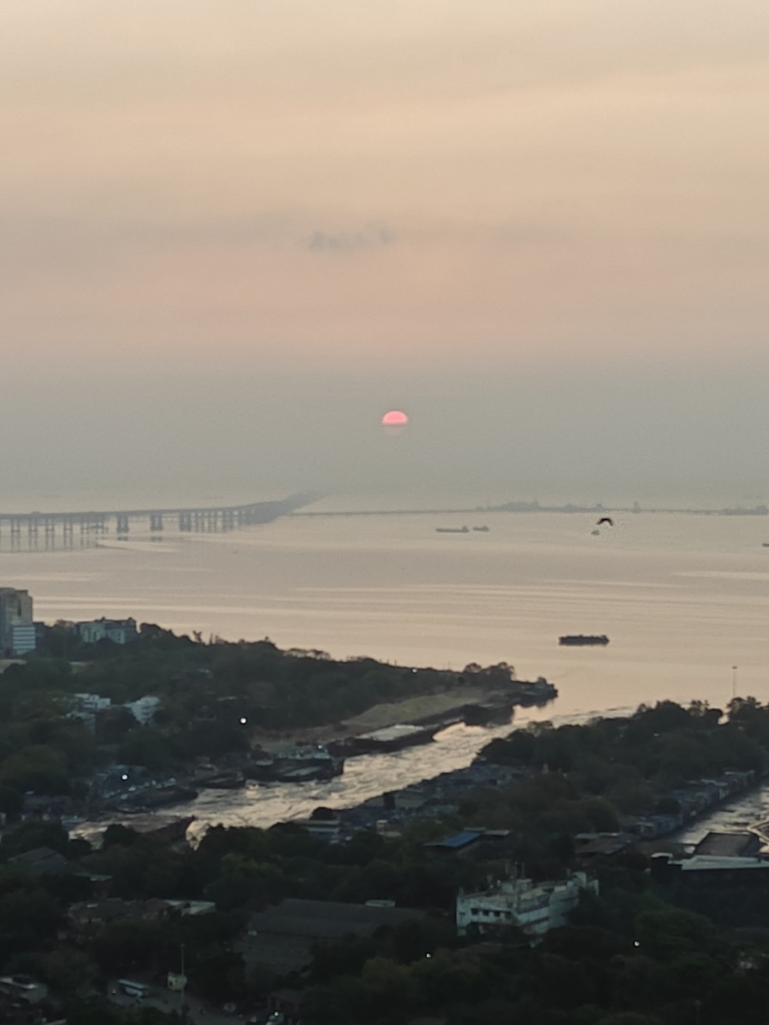The image shows a sunset view over Mumbai, India, likely from a high vantage point. The Bandra-Worli Sea Link bridge is visible spanning the water. The sun appears as a reddish disc near the horizon, obscured by haze. Several ships are visible on the water. In the foreground is a dense, urban landscape with buildings and vegetation.