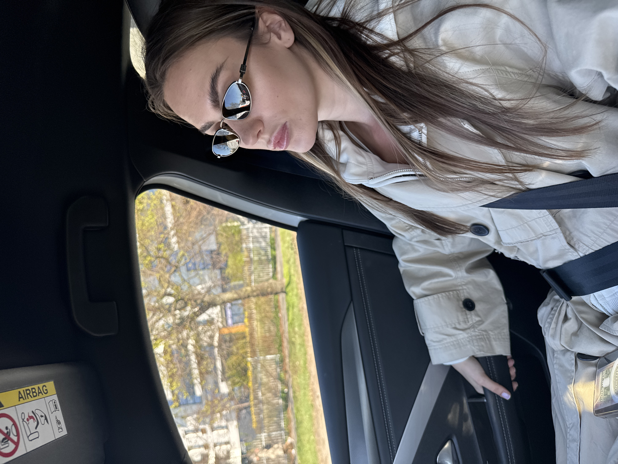 A young woman is sitting in the back seat of a car, wearing sunglasses and a beige trench coat. She is buckled in with a black seatbelt, and her hand is on the car door. The car appears to be moving, as a blurred view of trees and buildings is visible through the window. The trees indicate it is likely spring. The car's interior is black leather with silver trim. Based on the style of buildings seen through the car window, the location is likely the city of Warsaw, Poland. An "Airbag" warning sign is visible on the side of the car frame.