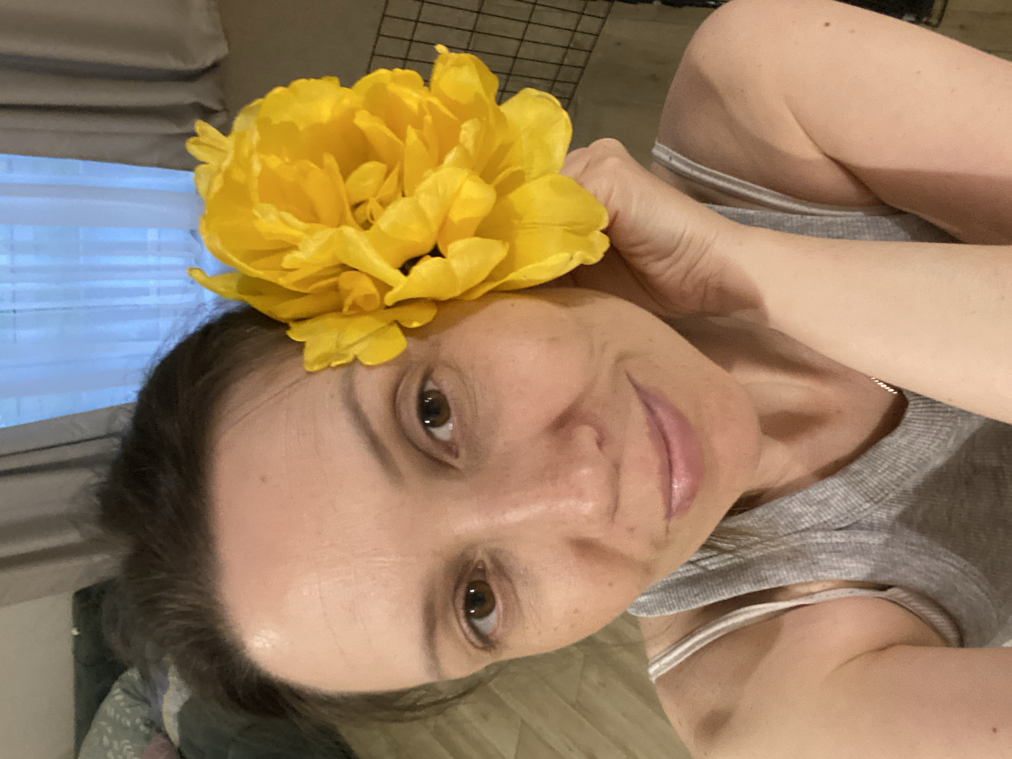 A woman is lying down, taking a selfie. She has a large yellow flower in her hair and her hand is near her face. She is wearing a gray tank top. The background includes a window with blinds and what appears to be a wire rack or cage. The location in Chaikovsky, Russia, is not discernible from the provided image.