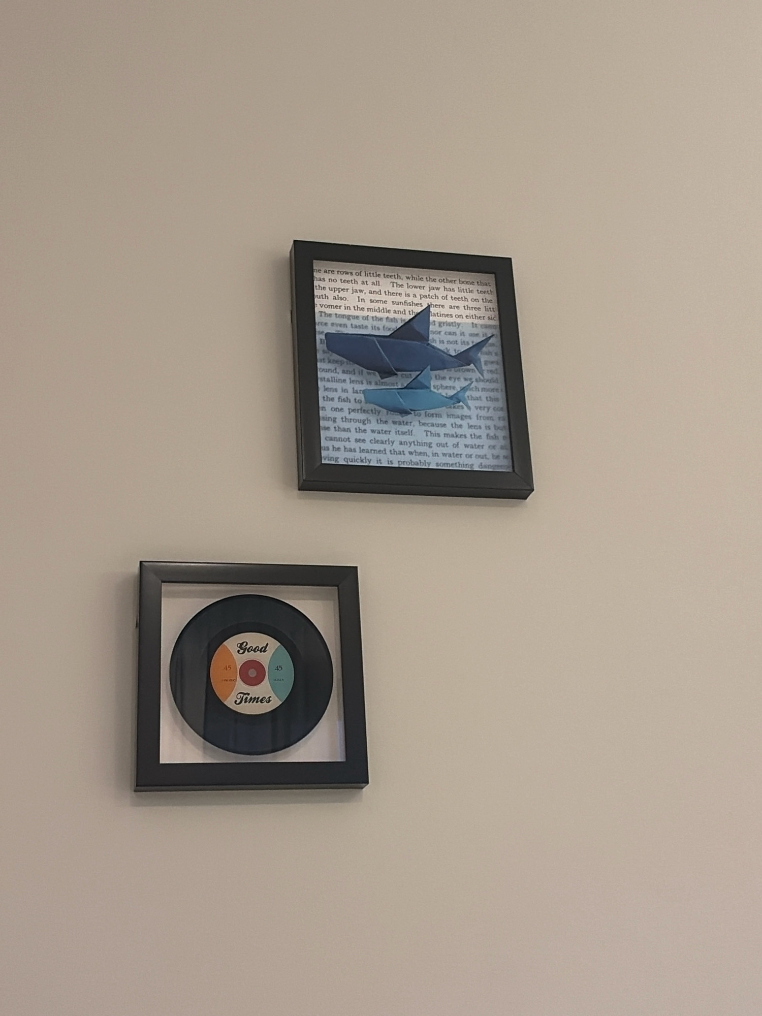 This is a photograph of two framed art pieces hanging on a plain, light-colored wall. The top frame contains an origami shark placed on pages of a book. The shark is a deep blue color and is folded in a way that resembles a sharp, predatory creature. The text on the book pages is mostly obscured by the shark and the framing, but appears to be printed in black ink. The bottom frame is square and contains a circular object that resembles a vinyl record. The record has colorful labels in the center with the words "Good Times" printed on it. There are also numbers "45" on the labels. The frames themselves are simple, dark rectangular borders. The lighting suggests an indoor environment, possibly a home or a small gallery. There is no indication of the location within Ras Al-Khaimah, United Arab Emirates.
