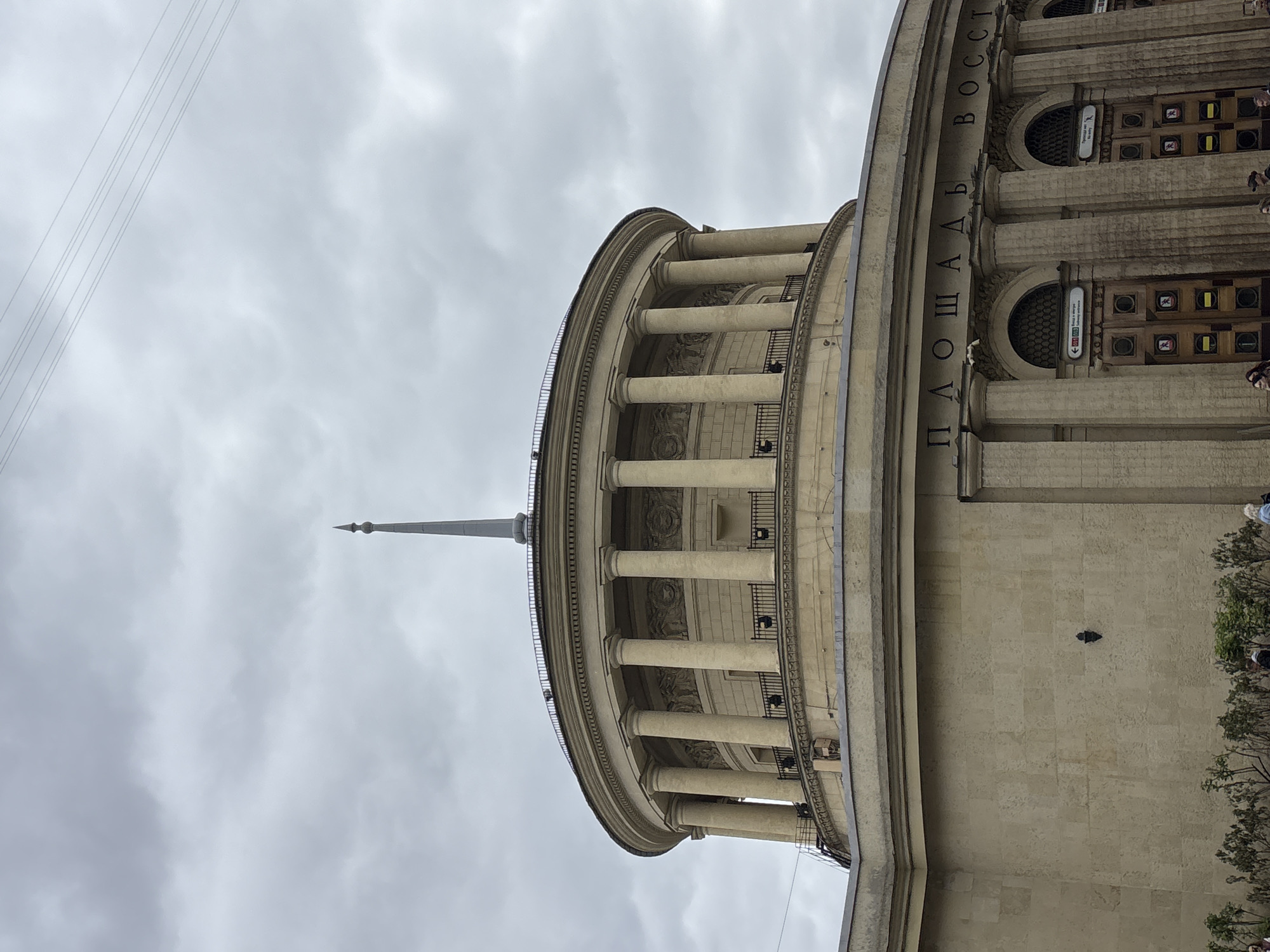 The image shows the upper part of the Moskovskaya metro station, located in Saint Petersburg, Russia. The view is from a low angle, capturing the station's rotunda adorned with classical columns and a spire. Vertical Russian text reading "Площадь Восстания" (Ploshchad Vosstaniya, meaning "Rebellion Square") is visible on the building's facade. The sky is overcast and gray. Power lines are also visible above the building. People and greenery are partially visible on the right side. A dark shape, probably a bird, is on the right side of the building.
