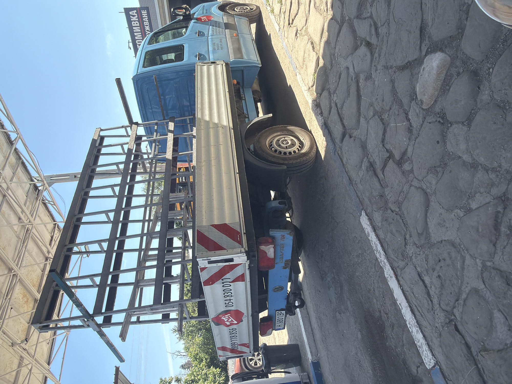 This image, captured in Shumen, Bulgaria, depicts a light blue flatbed truck with a large, intricate metal rack structure on its bed, likely designed for transporting glass panes or similar large, flat materials. The truck is parked on a cobblestone surface under a clear, bright blue sky, suggesting mid-day or early afternoon due to the strong sunlight and shadows.
The main flatbed truck displays a red "Zet" logo along with two phone numbers: "0899 118 856" and "054 830 017". Text on the cab side includes "СТЕКЛА" (Glass/Windows) and "ГОРНИ ДОБА". Its Bulgarian license plate is "H 8801 BP", with 'H' designating the Shumen region. The rear bumper also reads "GORN.FIBER". Behind it, another light blue van, possibly a Volkswagen, is partially visible.
In the background, a large sign is present, with the Cyrillic text "АВТОМИВКА" (Car Wash) and "ОБСЛУЖВАНЕ" (Service/Maintenance), indicating a vehicle service or repair establishment nearby. To the left, a metal scaffolding or a building frame is partially seen. The scene is functional and portrays commercial vehicles in an urban or industrial environment.