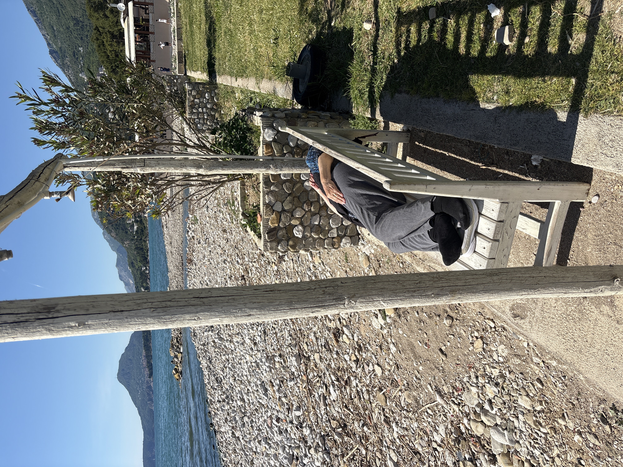 The image shows a person lying on a white wooden bench in Bar, Montenegro. The person is wearing grey clothing and black shoes, with a blue patterned head covering. The bench is situated between a rocky beach and a grassy area, with a stone wall nearby. In the background, there is a body of water and mountains under a clear blue sky. A wooden structure supports a tree adjacent to the bench. The scene is sunlit, casting shadows from the bench and nearby foliage.