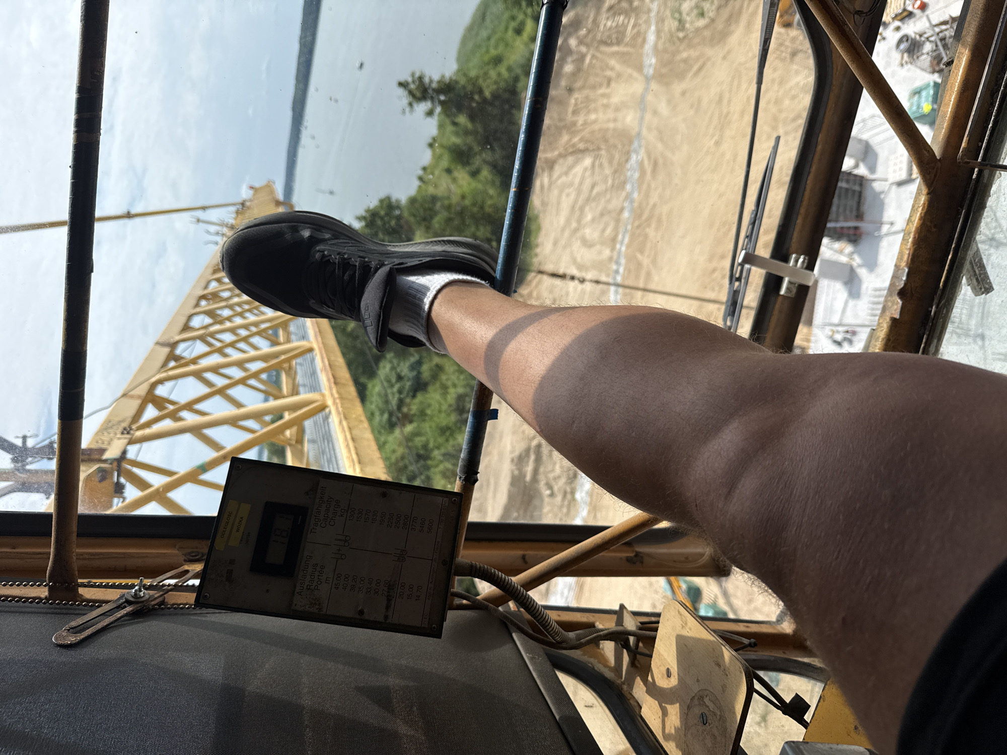 This image shows a first-person perspective from inside a crane cabin, looking out. The subjects are a person's leg and foot, wearing a black shoe and white socks, positioned on the edge of the cabin. The background reveals a construction site with a large yellow crane arm extending upwards and to the left. Beyond the construction site, a body of water, possibly a lake, is visible, with trees lining the far shore and a hazy sky above. The scene appears to be outdoors during the daytime, with sunlight casting shadows within the cabin.
A notable object in the foreground is a control panel or data plate with various measurements and labels. Text on this plate is in Polish and English, including "ODLEGŁOŚC WOZKA" (Trolley Distance), "Ausladung Radius Portée" (Outreach Radius), and "Tragfähigkeit Capacity Charge" (Load Capacity). Specific load capacities in kilograms are listed against corresponding outreach radii in meters. The time of day is likely late morning or early afternoon, given the bright sunlight. The weather appears to be clear. The location context of Mikołajki, Poland, suggests this crane is operating near a lake or waterfront area. The image conveys a sense of being high up and potentially involved in construction or maintenance work.