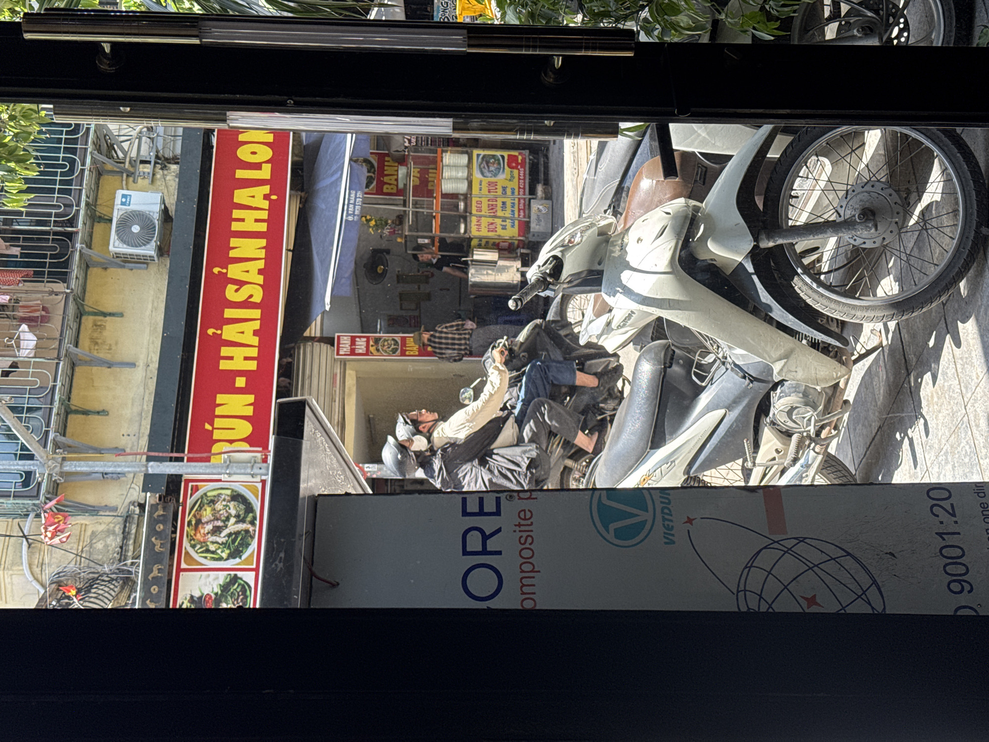 This image depicts a street scene in Hanoi, Vietnam, viewed through an open doorway or window. In the foreground, two people wearing helmets are riding a motorcycle. The person in the front is wearing a light-colored jacket and the person in the back is wearing a dark jacket. They are traveling on a street paved with concrete tiles, with several other parked motorcycles visible.
Behind the motorcycles, there is a storefront with a bright red sign featuring yellow text that reads "BÚN - HẢI SẢN HẠ LONG." There are images of food on the sign. To the left of the sign, there is a white panel with blue text and a circular logo featuring a globe. Further in the background, a food stall is visible with a vendor and customers. Several potted plants add greenery to the scene. The architecture suggests a typical urban setting in Vietnam, with buildings and awnings lining the street. The lighting indicates it is daytime, likely sunny, casting shadows on the ground.