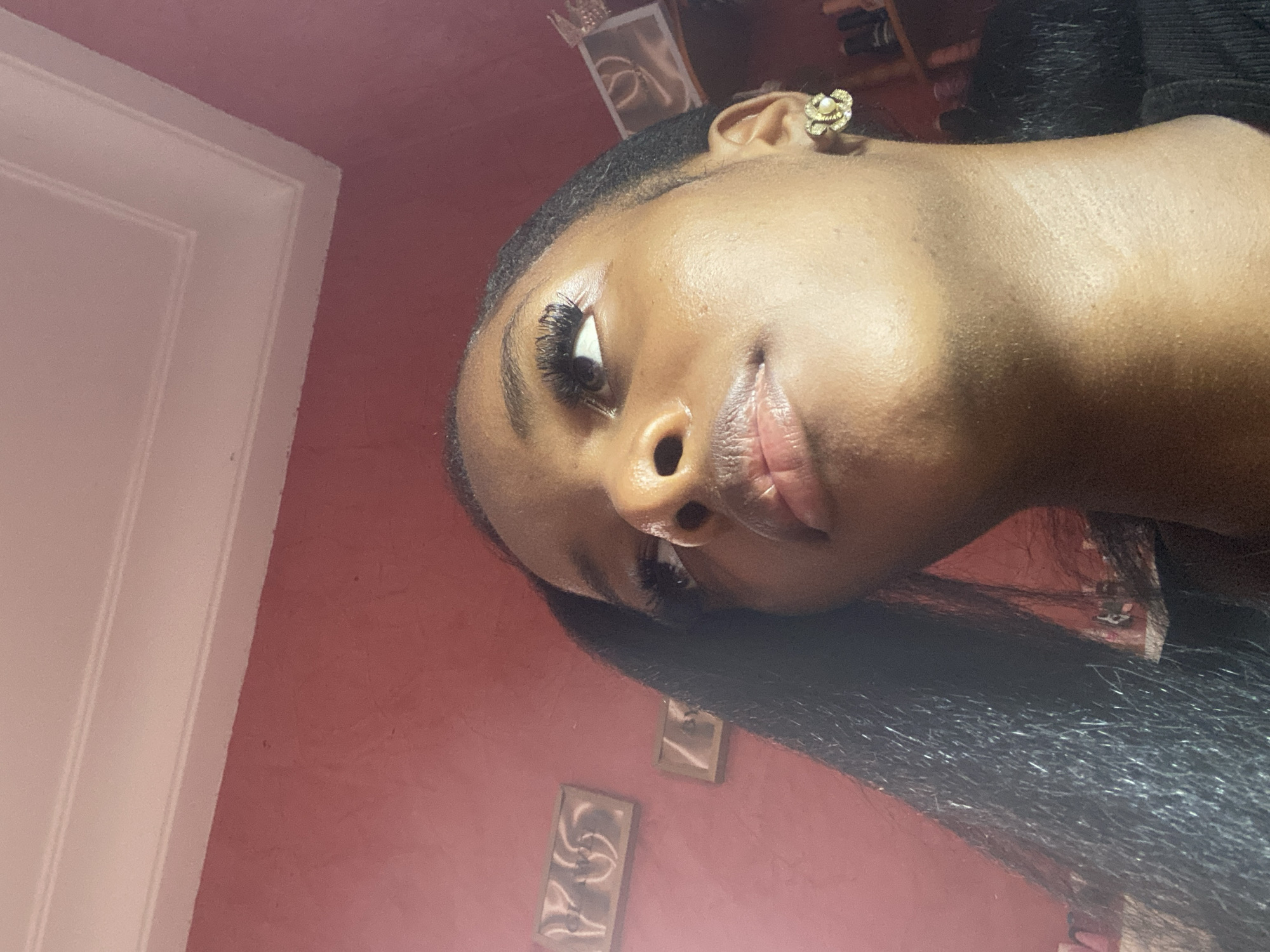 A low-angle close-up shot focuses on a young Black woman's face, looking upwards and to the left. Her right ear is adorned with a gold flower-shaped earring featuring a pearl in the center. Her dark hair cascades down the left side of the frame. The background consists of a reddish-pink wall with decorative trim at the top, and two framed pieces of art on the wall to the left. On the right, shelves with various items are partially visible. The overall impression is an indoor setting, possibly a home in the city of Jos, Nigeria.