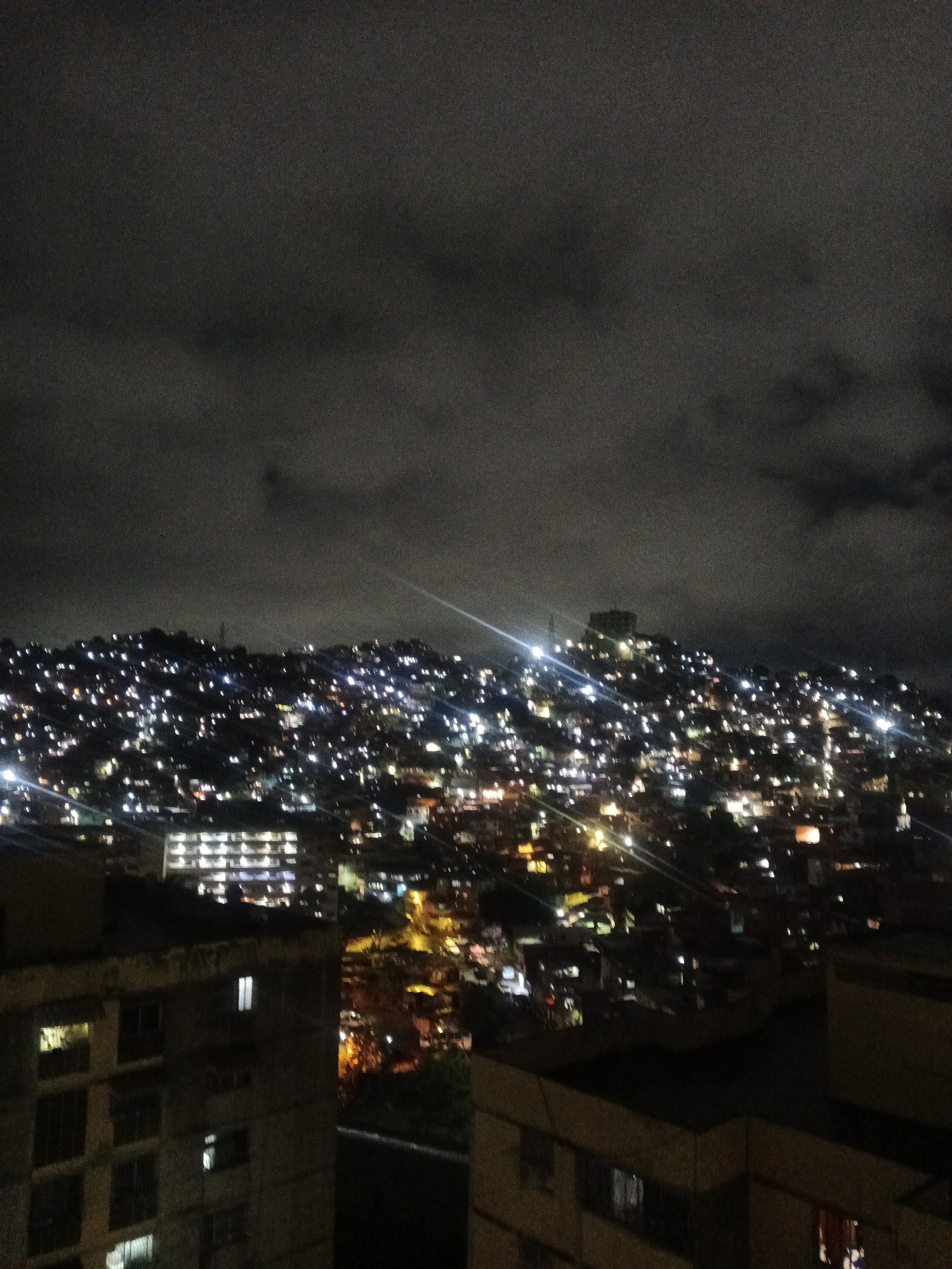 The image shows a nighttime aerial view of a densely populated hillside in Caracas, Venezuela. Numerous lights from buildings and streets illuminate the dark landscape, creating a sparkling effect. The sky is cloudy and dark. In the foreground, there are several buildings, some with visible windows lit from within. The hillside itself is covered with structures, suggesting a favela or informal settlement, common in Caracas. The overall impression is one of a vast, sprawling urban area at night, characterized by its density and the pervasive glow of artificial light.