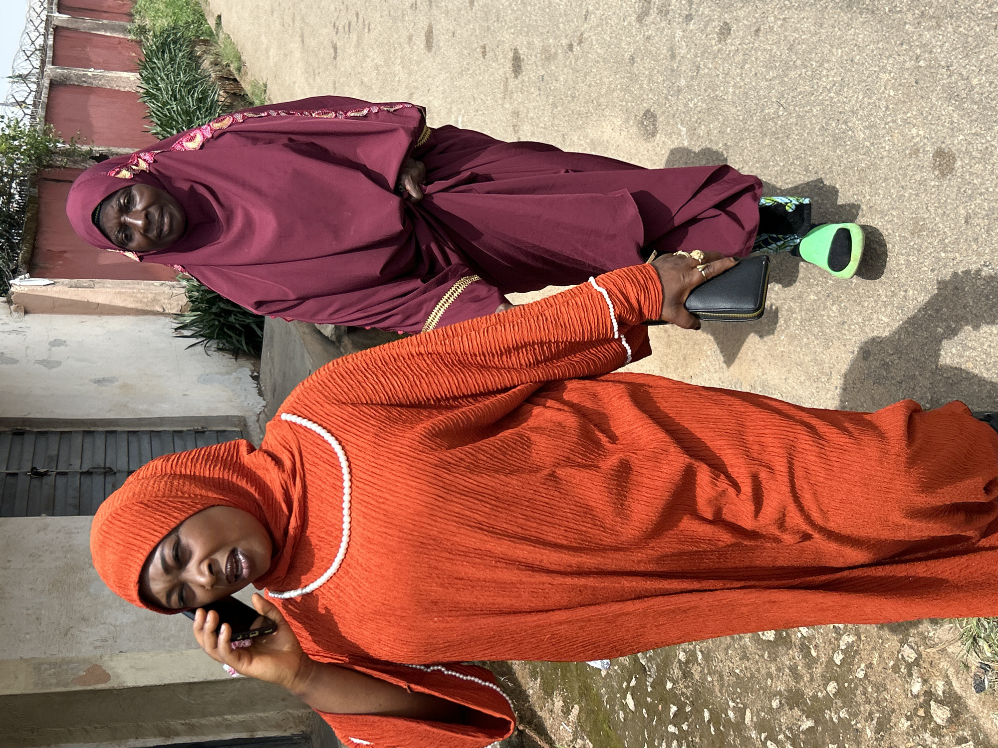 Two women are standing on a paved surface next to a building and some vegetation. One woman is wearing an orange garment and a head covering, and is talking on a cell phone. She is also holding a black wallet or small purse. The other woman is wearing a dark burgundy garment and head covering with floral embellishments. A green object is lying on the ground next to them. The location is suspected to be Jos, Nigeria.