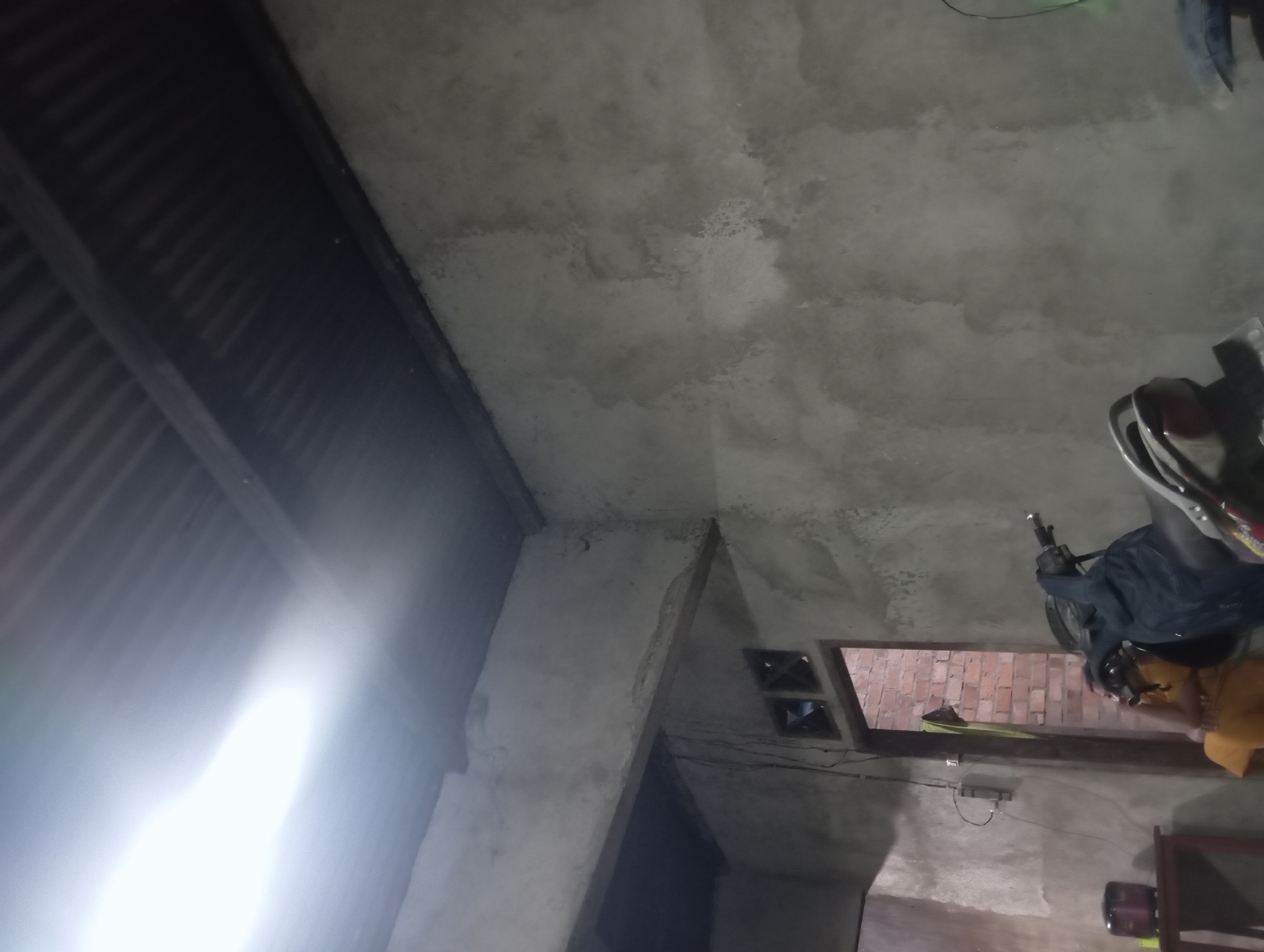 The image depicts an interior scene, likely a garage or workshop, with a corrugated metal roof visible at the top. The walls are unplastered concrete, showing a rough texture. A bright light source is prominent in the upper left corner, casting a strong glare. In the foreground, a dark-colored motorcycle is parked, with a portion of its handlebars and seat visible. To the left of the motorcycle, a wooden table with a dark-colored appliance on it can be seen. A doorway is visible in the mid-ground, leading to an area with exposed brick walls. A person, partially obscured, is seated in the doorway, wearing a yellow garment. The overall impression is of a functional, unadorned space, potentially in a rural or less developed area, consistent with many structures in Rejosari, Indonesia.