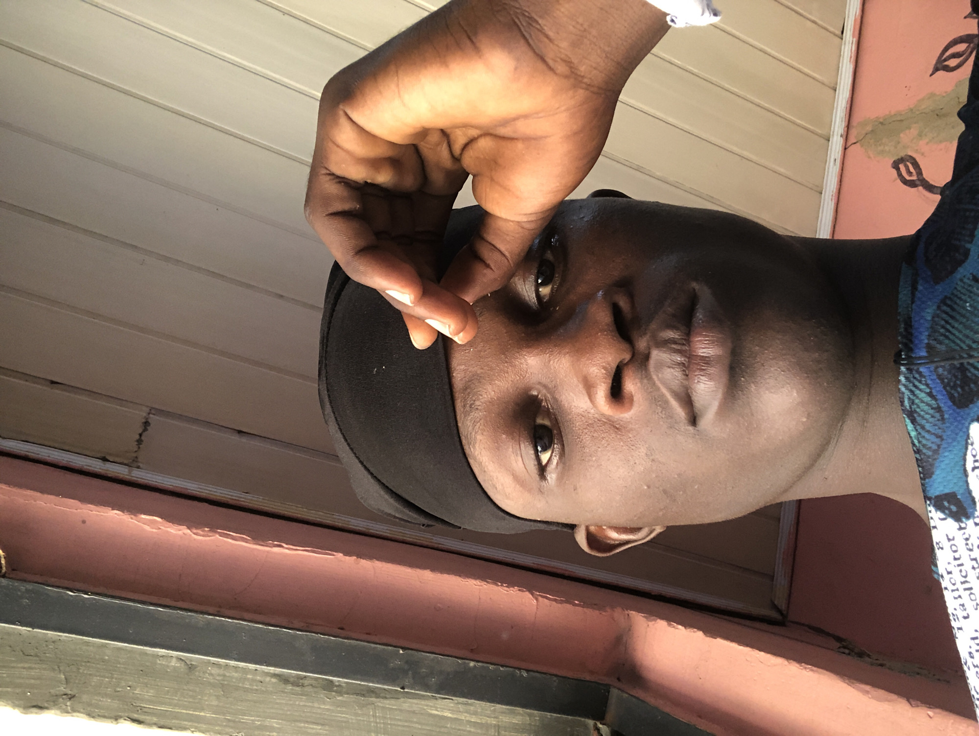 A low-angle, close-up, outdoor selfie shows a young Black man from the chest up. He is wearing a black beanie and a patterned blue and black shirt. His right hand is raised, with his index finger and thumb touching his forehead. He is looking directly at the camera, with his eyes slightly narrowed. The background consists of a pink wall and a wooden ceiling. The scene is likely from Ado, Nigeria.