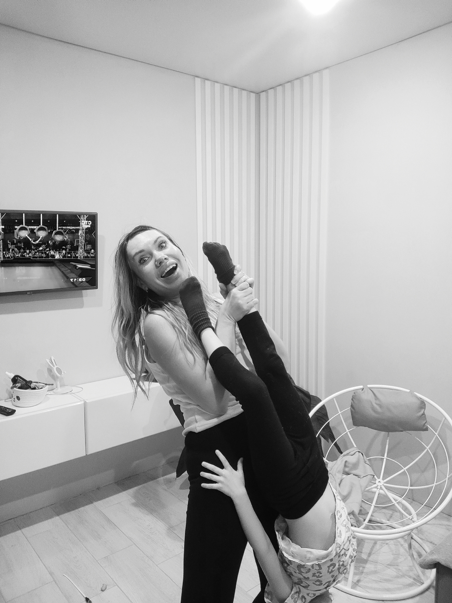 The photograph, taken indoors, depicts a woman holding a child upside down. The woman has long hair, is wearing a light-colored top and dark pants, and is smiling with an open mouth. The child, dressed in dark leggings and a light-colored patterned shirt, is being held by their feet.
In the background, there's a TV screen displaying a scene of people on a water obstacle course. Beneath the TV is a white cabinet with a basket and other items on it. A white wire-frame chair with a cushion is positioned to the right. The walls are a light color, with a section of vertical stripes.
The setting is a modern interior space, potentially a living room. There's no indication that this is Votkinsk, Russia, or any city in Russia.