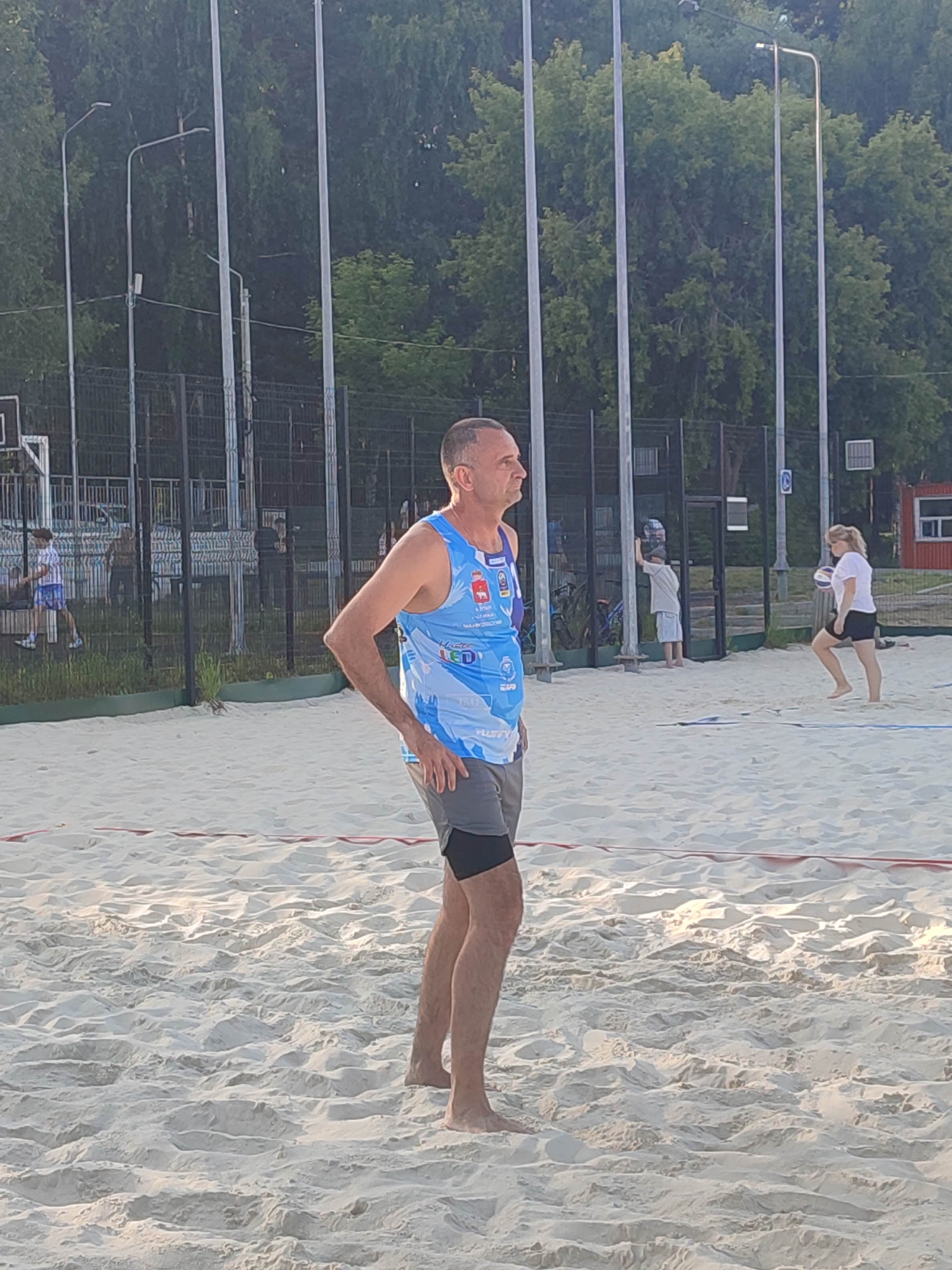 An outdoor beach volleyball court is depicted. A man, wearing a blue and white sleeveless shirt and grey shorts, stands in the foreground, facing right, with his hands on his hips. His left arm is bent at the elbow, and his right hand is resting on his hip. The sand is uneven and textured. In the background, several tall light poles with lamps are visible. Behind the poles, a chain-link fence encloses the court. Beyond the fence, there are trees. A woman in a white t-shirt and black shorts is visible on the court in the middle ground, holding a volleyball. Another person, possibly a child in a light-colored t-shirt and shorts, stands near the fence. A red structure with a white window is present on the far right of the background. The overall scene is brightly lit, suggesting daylight.