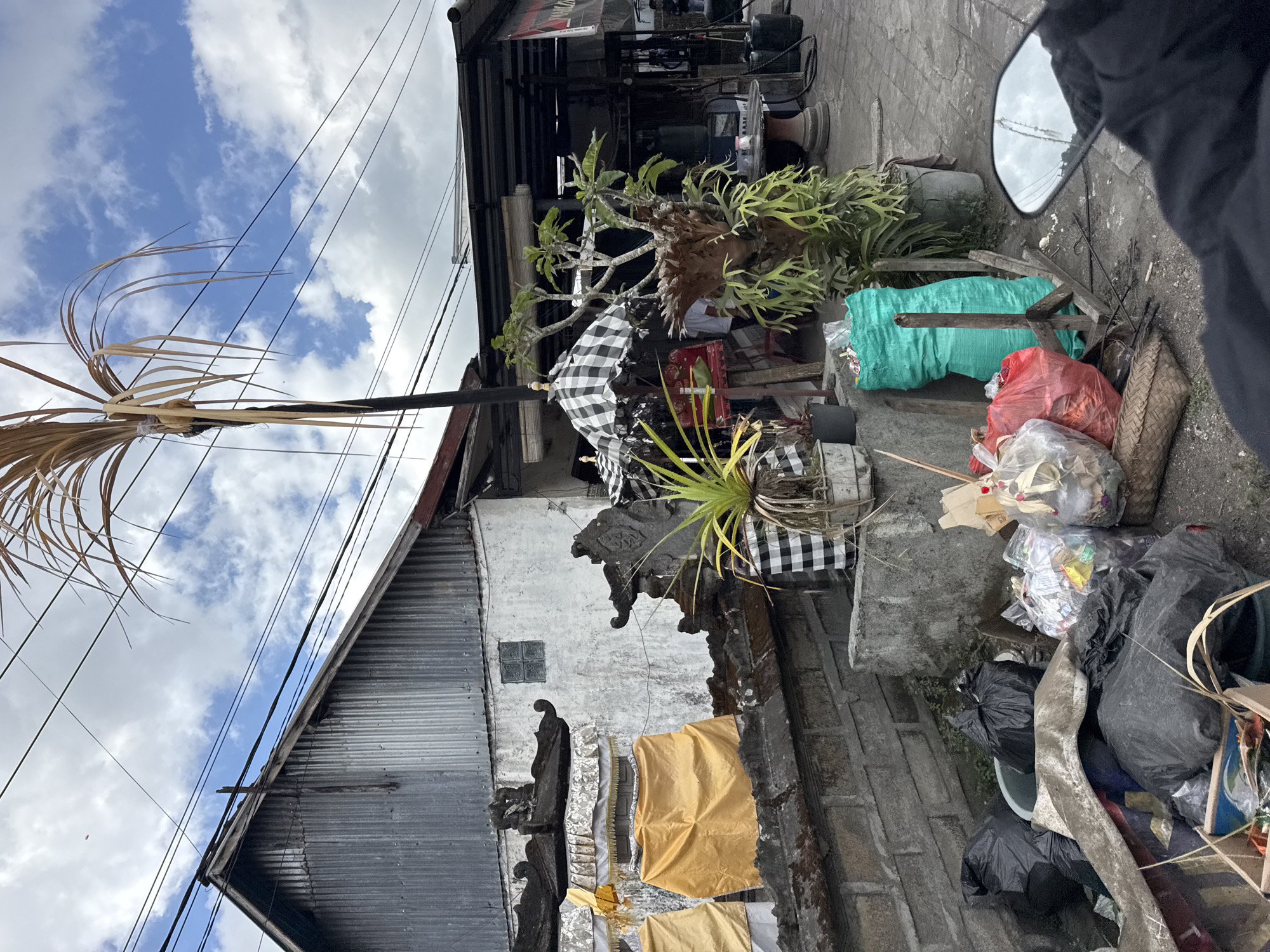 This image depicts a street scene in Kintamani, Indonesia, characterized by a collection of trash bags and debris piled on the roadside. In the foreground, several black trash bags, a blue basin, and a woven basket are visible. Behind these, a large green refuse sack, red trash bags, and other assorted garbage are scattered.
The background features traditional Balinese structures with ornate stone carvings, adorned with yellow and white fabric coverings. A tin-roofed building with a white plastered wall stands to the left. Further back, a covered outdoor area with tables and chairs suggests a commercial establishment, possibly a shop or restaurant. A large plant with wide leaves and a dried palm frond structure are prominent elements of the scene. The sky above is partly cloudy, with visible electrical wires crisscrossing the frame. The perspective suggests the image was taken from a moving vehicle, possibly a motorcycle, as indicated by a rearview mirror in the lower right corner. No people are clearly discernible in the scene.