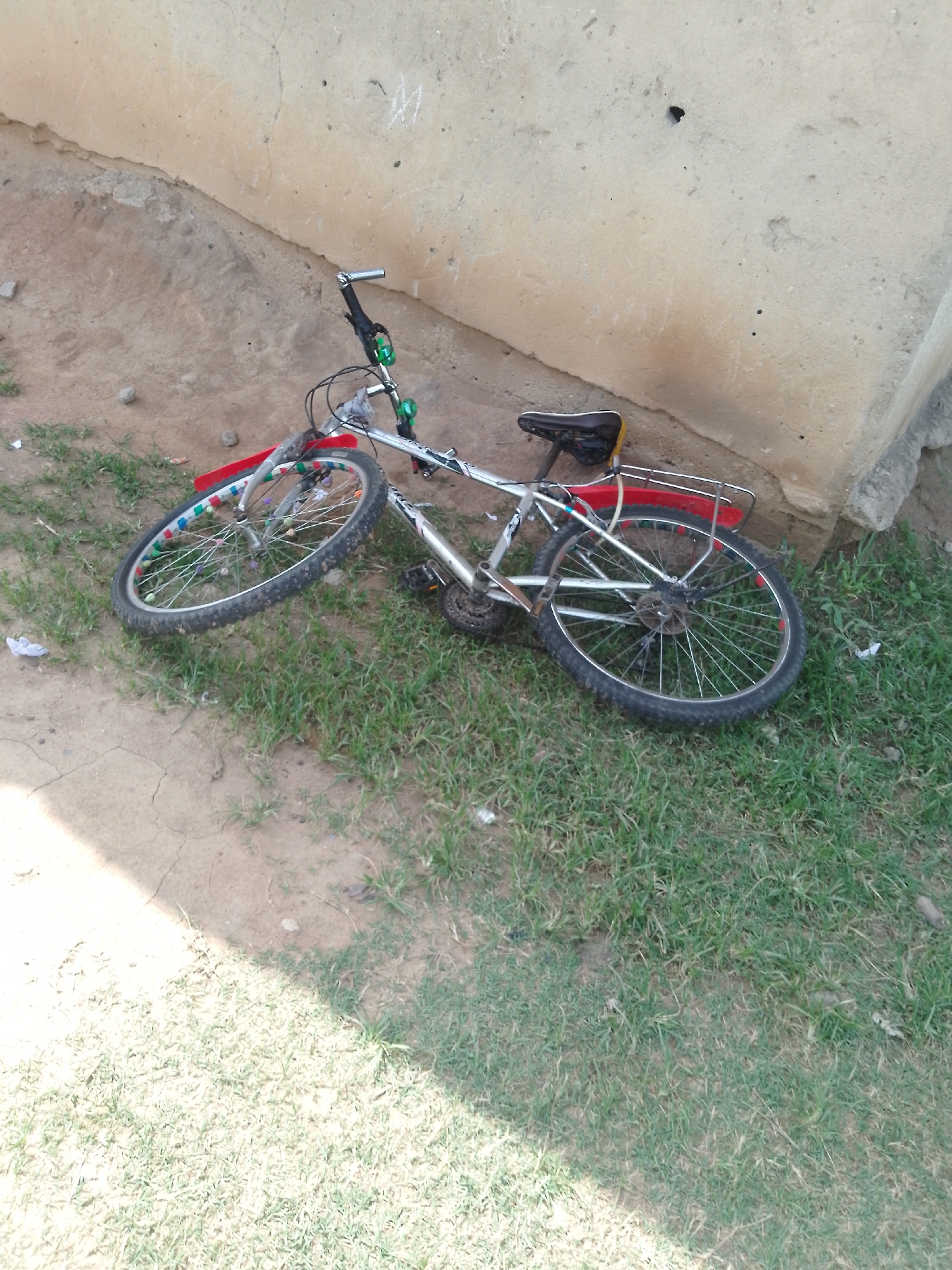 The image shows a silver bicycle with a black seat, resting against a textured, light-colored wall. The bicycle has a red rear fender and colorful decorations on its spokes. The front wheel is turned to the left, and the bicycle is lying on its side on a patch of grass and dirt. The background consists of a cracked, sandy ground and a rough, beige wall, possibly part of a building. The overall scene appears to be outdoors, in what could be an urban or semi-urban environment. There are no people or animals visible in the image. The location is identified as Serti, Nigeria.