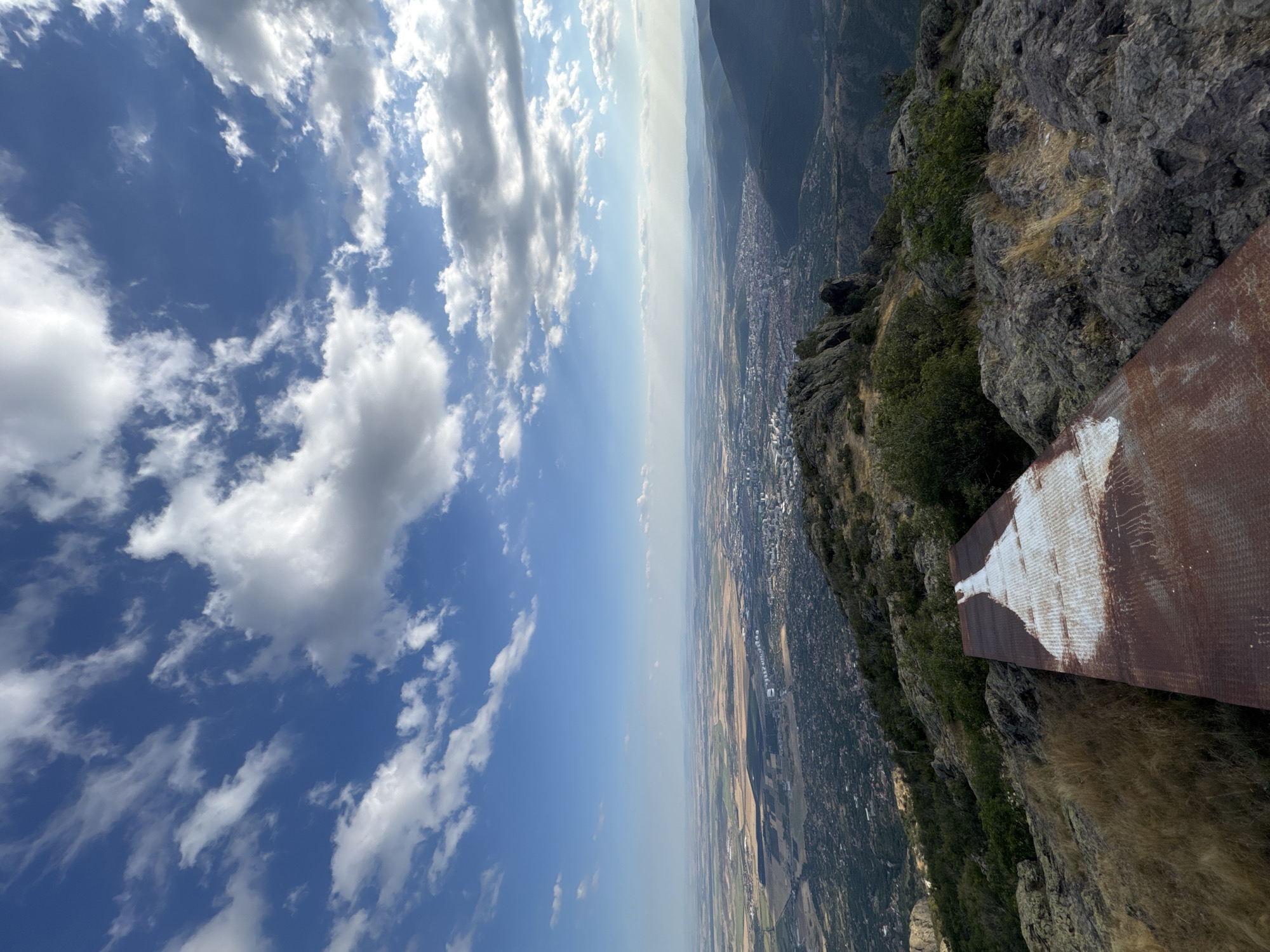 The image is a landscape photograph taken from a high vantage point, looking out over a city and surrounding countryside. In the foreground, a rusted metal platform with a white, painted shape on it is angled towards the left. The platform rests on rocky terrain with dry grass.
Beyond the immediate foreground, the terrain slopes downward, revealing a city nestled in a valley. The city appears to be densely populated with buildings. The countryside surrounding the city consists of fields, some appearing cultivated with shades of brown and green, and hills in the distance.
The sky is a vibrant blue, dotted with white, fluffy clouds. The lighting suggests it is daytime, with the sun casting shadows and highlighting certain areas. The overall scene depicts a panoramic view of an urban area and its natural surroundings from a mountainous or elevated position. This view is consistent with the location of the Cherni Vrah peak, near the city of Ihtiman, Bulgaria.