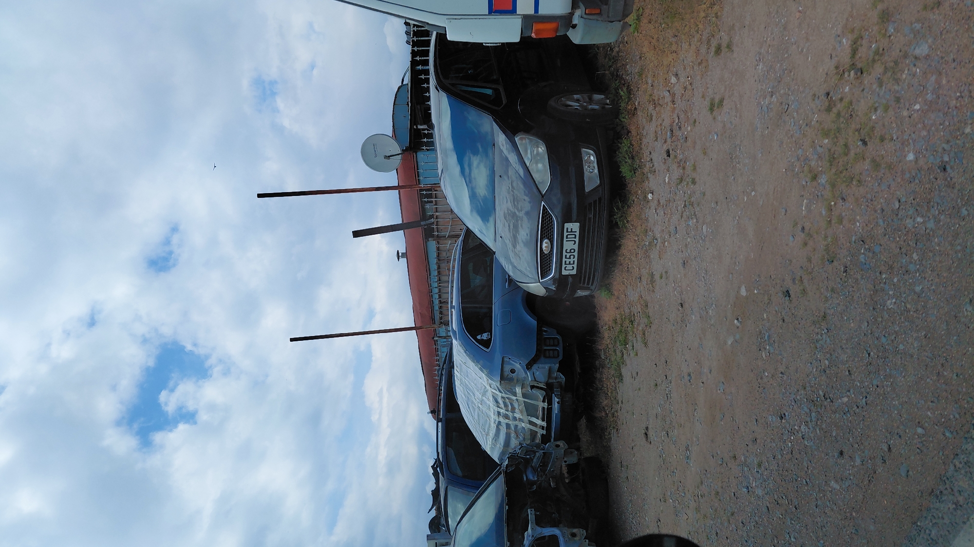 The image depicts a junkyard or vehicle dismantling facility. The foreground shows multiple partially dismantled cars, primarily blue in color. Car bodies are stripped of parts, with some covered in plastic sheeting. In the mid-ground, a black car with license plate "CE56 JDF" is visible. A white van with a red and blue stripe is situated towards the back. The ground is covered in dirt and gravel. The background features a cloudy sky, with a structure partially visible, including what appears to be a satellite dish. The location is given as Unknown, Unknown.
