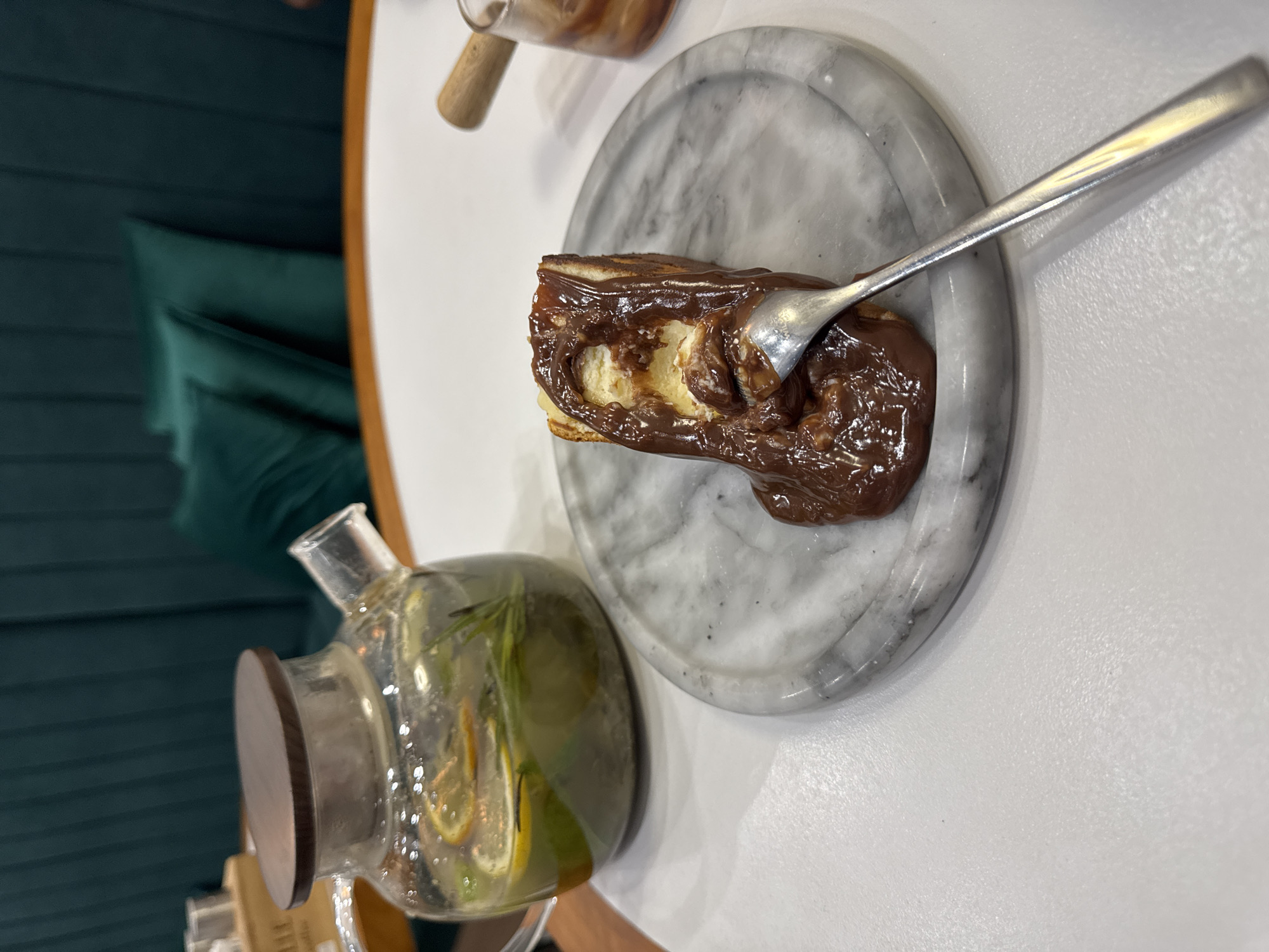 An overhead shot shows a slice of cheesecake on a marble plate, topped with melted chocolate. A fork is embedded in the cheesecake, seemingly mid-bite. To the left, a glass teapot with a wooden lid contains a clear liquid with slices of lemon and sprigs of rosemary. The scene is set on a white table, with a dark green upholstered booth visible in the background. The image appears to have been taken in a cafe in Bishkek, Kyrgyzstan, indicated by the presence of common cafe elements such as a teapot and dessert.