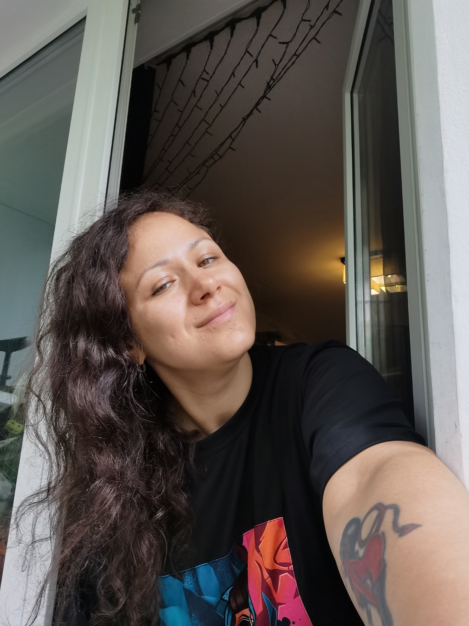 A woman with long, dark, wavy hair and light brown eyes is depicted in a close-up photograph, looking directly at the camera with a gentle, slight smile. She appears to be leaning out of or standing beside an open white window or door frame. She is wearing a black t-shirt that features a colorful graphic design with shades of blue, orange, red, and pink on its front. A prominent tattoo, illustrating a stylized figure with a tail and a red, heart-like shape, is visible on her left upper arm.
The background inside the room is dimly lit, showcasing string lights (fairy lights) draped across the ceiling and a visible ceiling light fixture reflected in the window pane. The lighting on her face suggests a bright, possibly natural, light source from outside the frame, contrasting with the darker interior. The scene captures a casual moment, likely within a home in Chaikovsky, Russia. Her expression conveys a sense of ease and friendliness. There is no discernible text visible in the image.