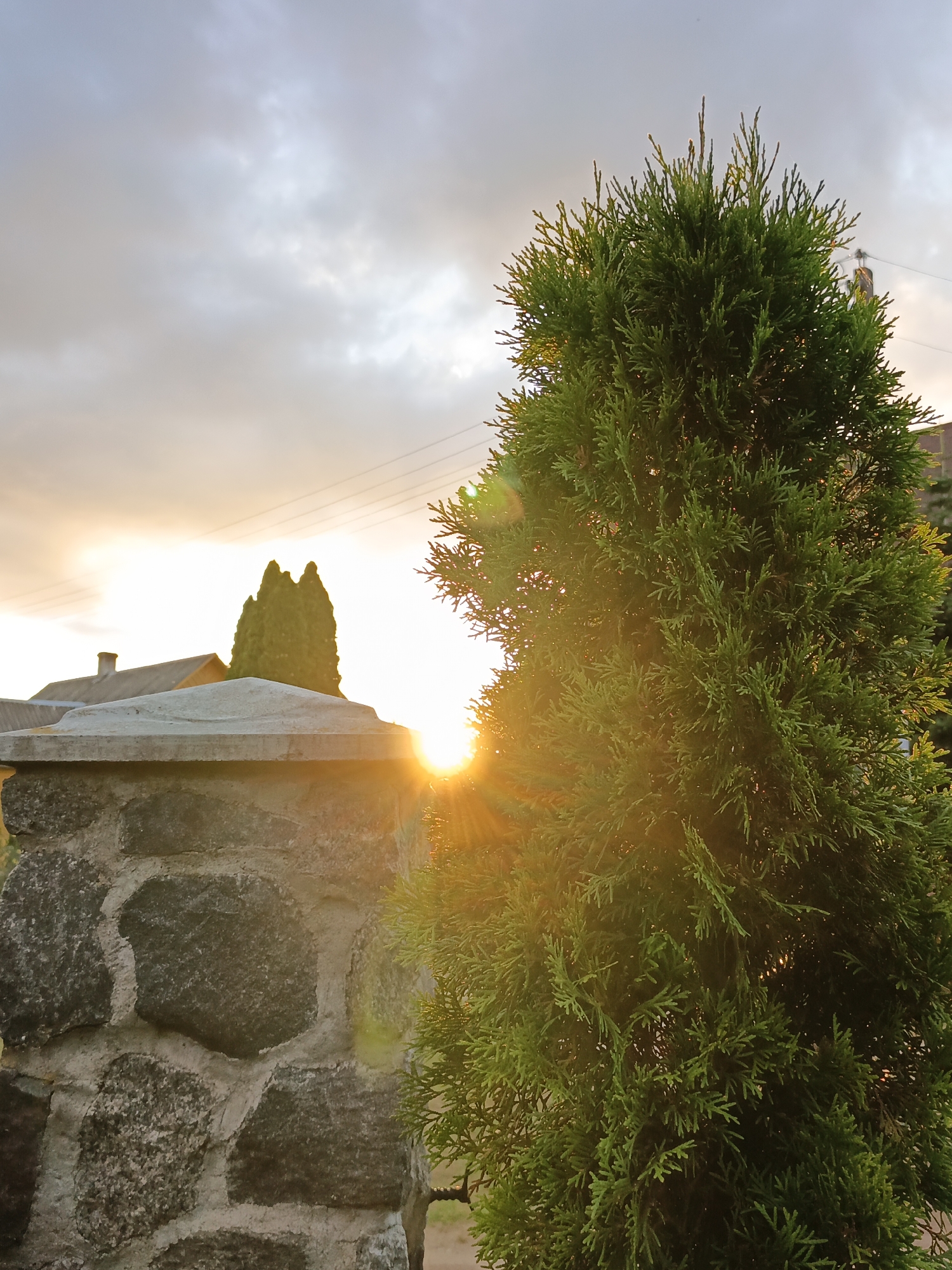 The image shows an outdoor scene in Лосева, Belarus. In the foreground to the left is a stone pillar made of rough-hewn, grey stones mortared together. To the right and slightly behind the pillar is a dense, green coniferous tree. The sun is low in the sky, creating a warm glow and sun flares that illuminate the tree. In the background, there is a residential building with a visible chimney and a sloped roof. Beyond that, there are power lines stretching across the sky, which is filled with clouds tinged with the orange and yellow hues of sunset. The overall impression is a peaceful evening in a rural or suburban setting. There are no people visible in the image.