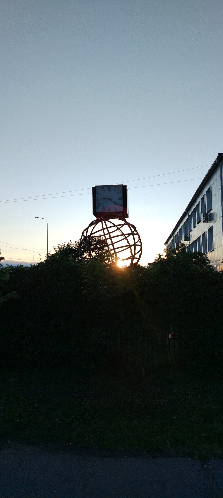 The image depicts an outdoor scene in Chaikovsky, Russia, at either sunrise or sunset. A prominent monument dominates the center, featuring a large, square clock face with Roman numerals, indicating approximately 10:45 or 10:50. This clock sits atop a spherical, open-cage metal structure, resembling a globe, which is partially obscured by dense, dark green foliage.
The sun is positioned low behind the base of the spherical structure, casting a bright, radiant glow through its framework and silhouetting the monument and surrounding bushes. The sky above is clear and a pale blue, transitioning to a warmer tone near the horizon.
To the right, a long, multi-story building with a light-colored facade and numerous windows is visible, reflecting some of the sky's light. To the left, a street light pole is silhouetted against the bright background, with faint power lines stretching across the upper portion of the sky. The foreground shows a dark, paved surface, likely a road or path, adjacent to a strip of grass. The overall atmosphere is serene and tranquil. No people are visible.