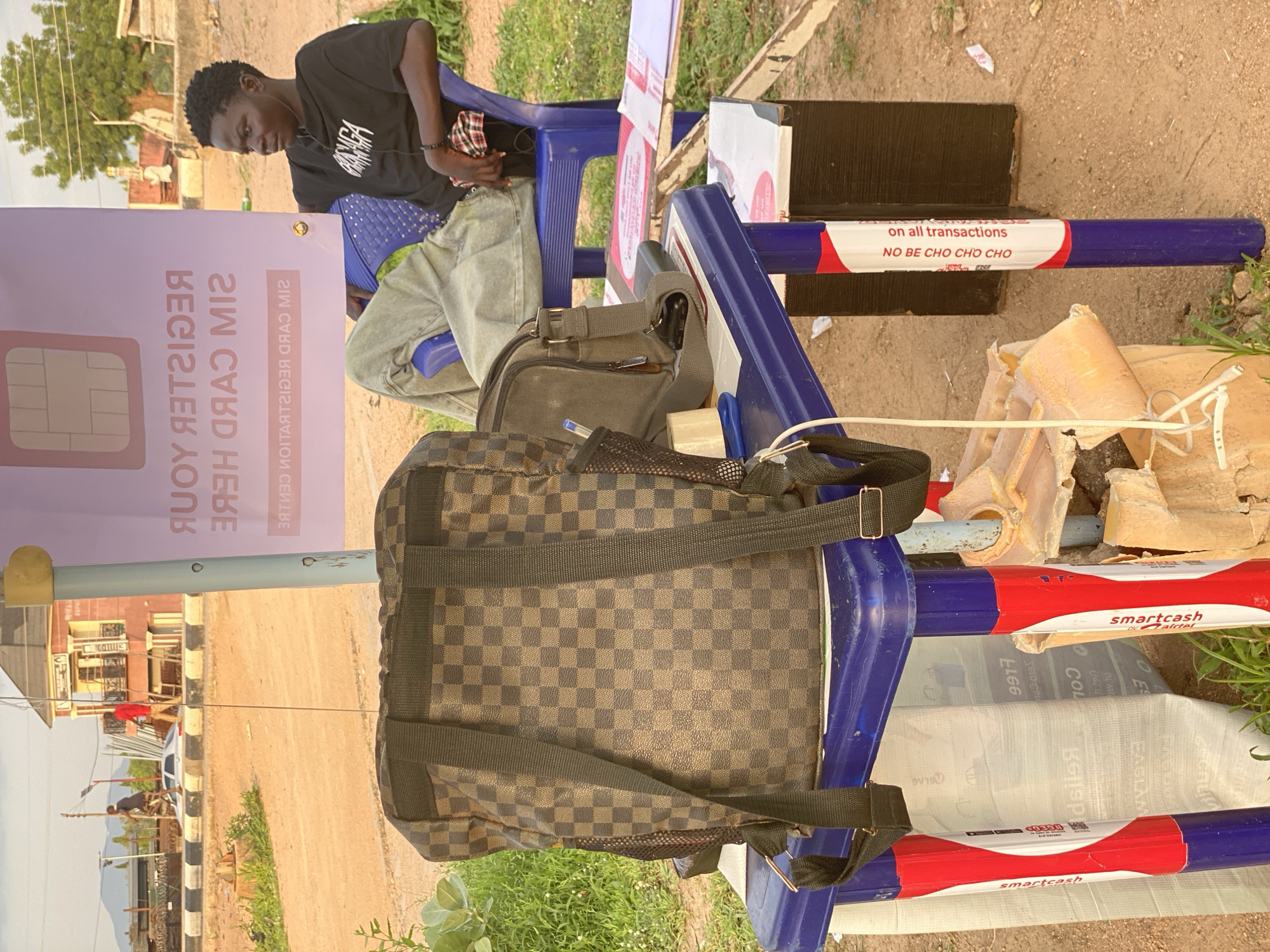 In Jalingo, Nigeria, a young Black male is seated on a blue plastic chair. He is wearing a black t-shirt with white lettering and light-colored pants. He has earbuds in and is looking to his right. Beside him, a blue plastic table holds a brown and black checkered backpack and a dark gray messenger bag. Attached to the table legs are red and white stickers that read "smartcash" and "NO BE CHO CHO CHỌ". Behind the table and chair, a purple banner with a SIM card graphic is visible. The background shows a dirt road, some buildings, and vegetation.