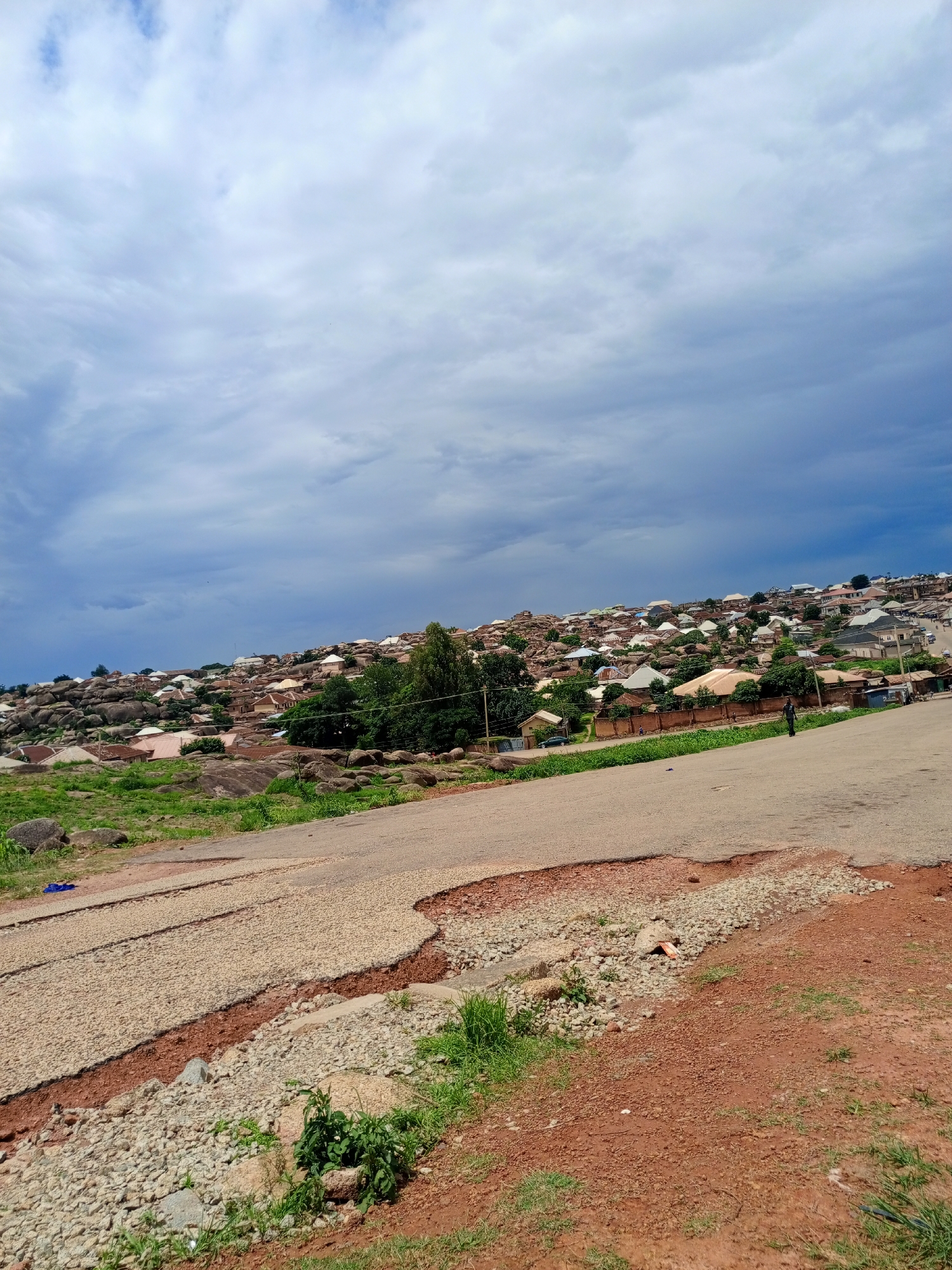 This is a photograph of a hillside residential area in Jos, Nigeria, under a cloudy sky. The foreground features a damaged asphalt road with exposed gravel and red soil. The road appears to be undergoing repair or has deteriorated. In the midground and background, a densely populated settlement covers the rocky incline. Numerous houses, characterized by their varied roofing colors including white, orange, and brown, are nestled amongst large boulders and green vegetation. The architecture suggests a blend of modern and traditional construction. The overall impression is of a community built on a challenging, uneven terrain, with signs of ongoing infrastructure work in the foreground.