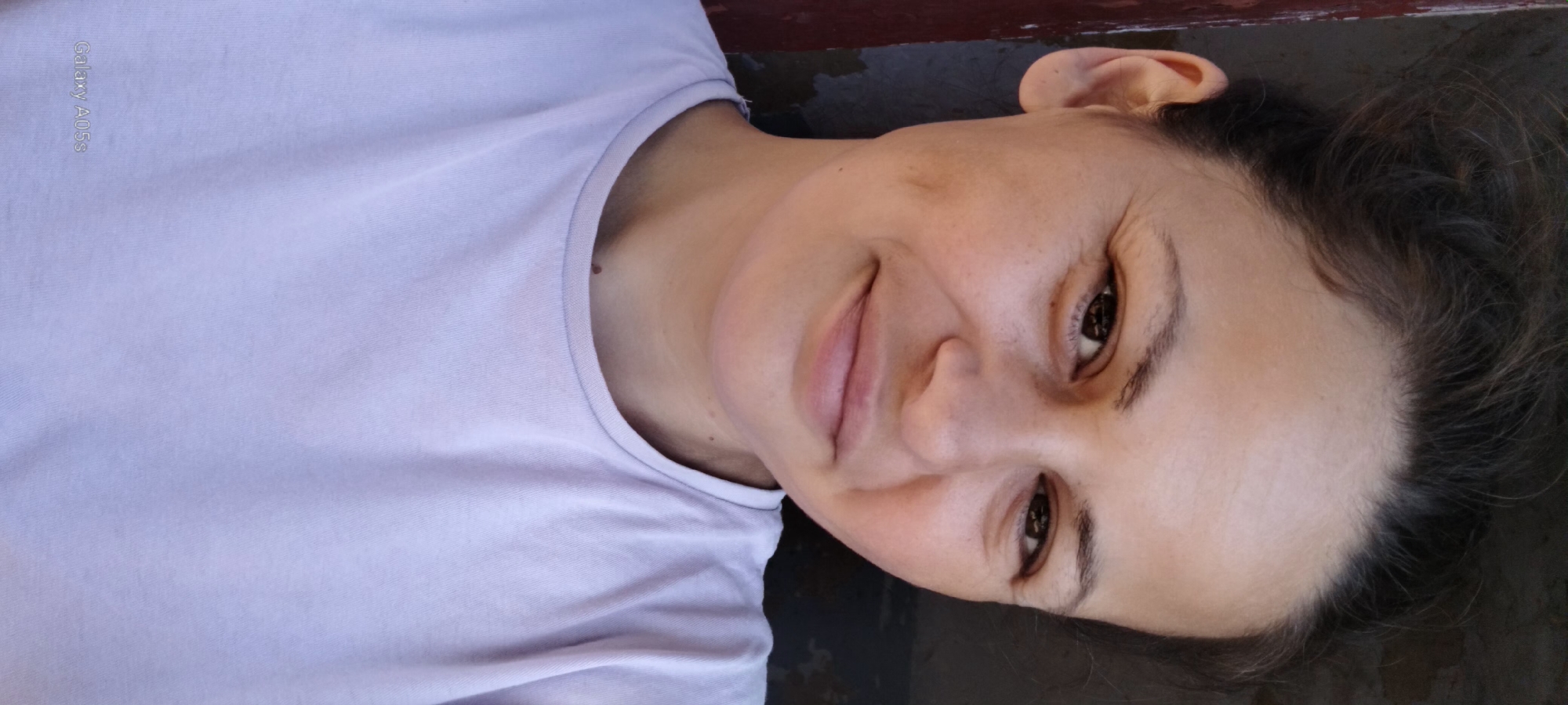 A close-up, shoulder-up, selfie-style portrait of a woman with brown hair pulled back and brown eyes. She has fair skin with a few freckles and dimples on her cheeks. She is wearing a light purple t-shirt with a crew neck. The background is out of focus, showing a dark wall and a reddish-brown vertical surface on the left. The lighting appears to be natural daylight. A watermark reading "Galaxy A05s" is visible in the bottom left corner.